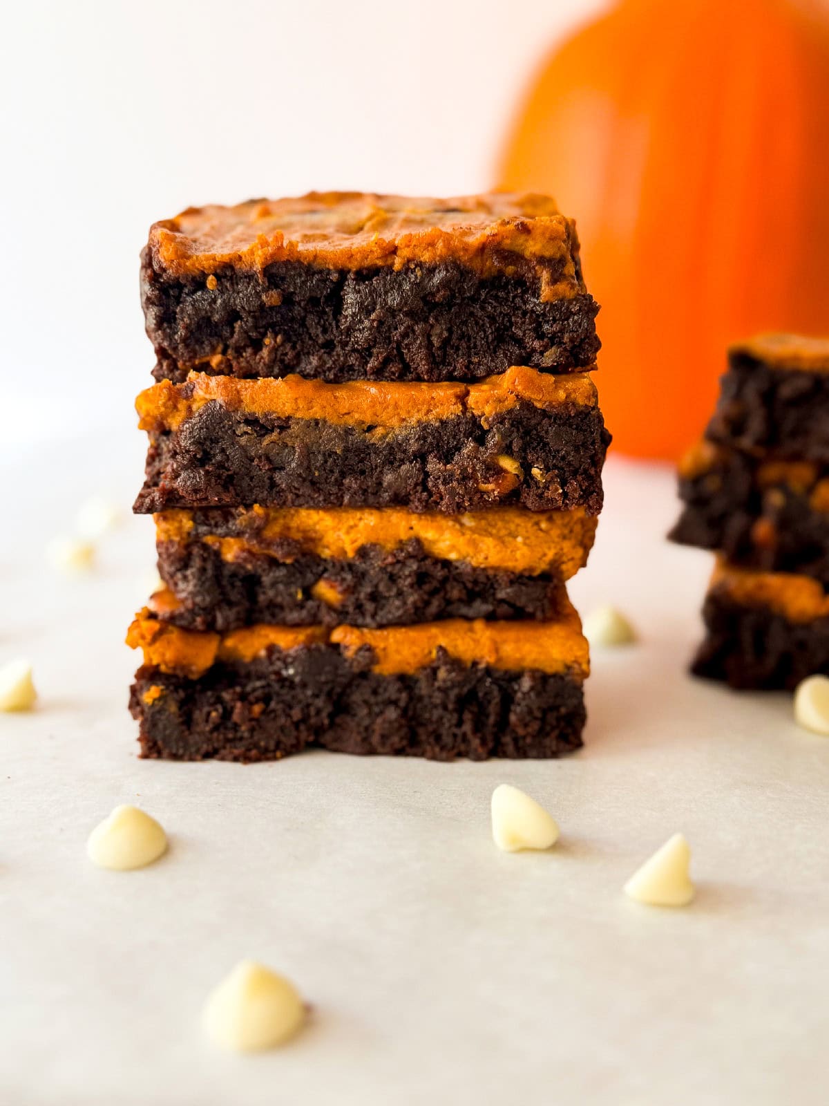 Tall stack of pumpkin cheesecake brownies highlighting the swirled chocolate and pumpkin layers on parchment paper with white chocolate chips and a blurred pumpkin behind.