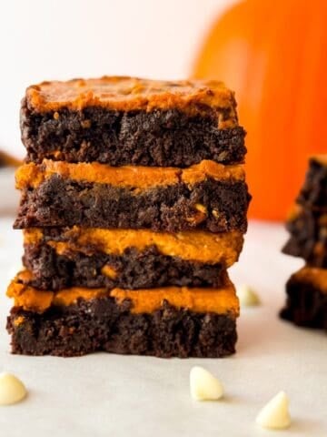 Stack of homemade pumpkin cheesecake brownies with fudgy chocolate layers, surrounded by white chocolate chips with a blurred pumpkin in the background.