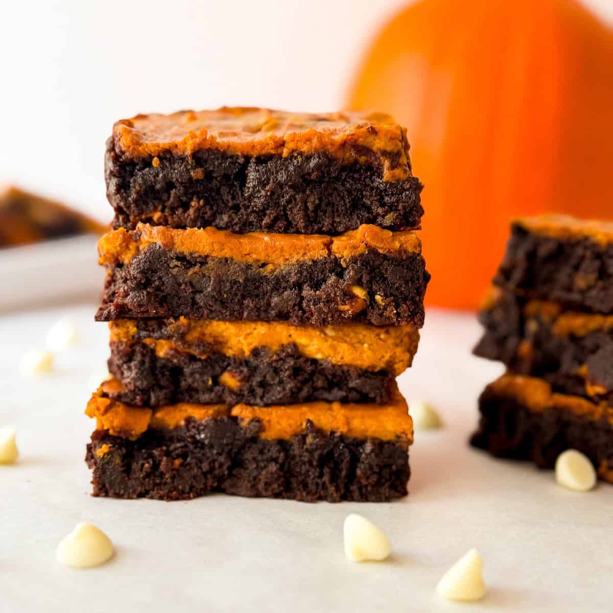 Stack of homemade pumpkin cheesecake brownies with fudgy chocolate layers, surrounded by white chocolate chips with a blurred pumpkin in the background.
