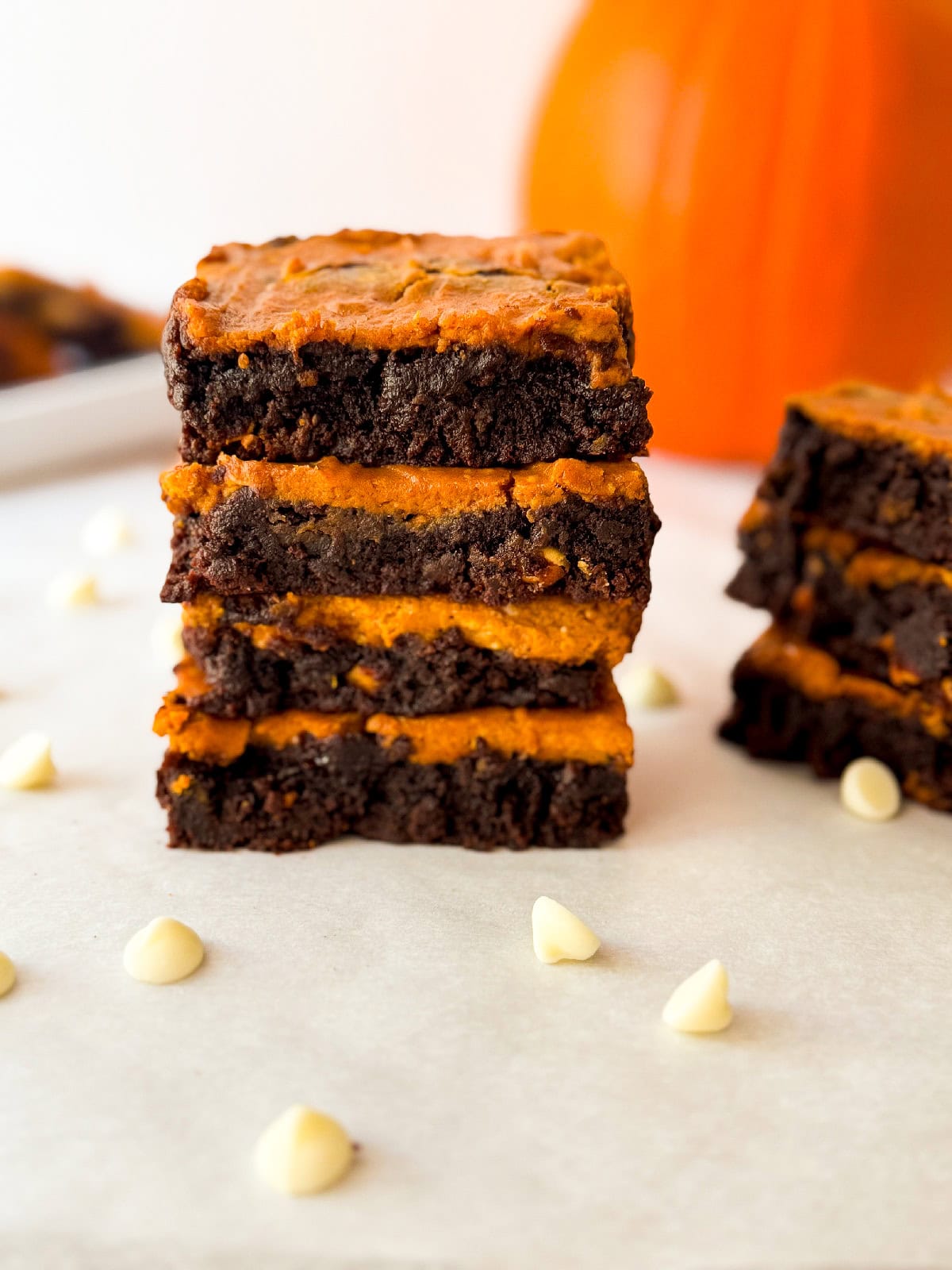 Stack of pumpkin cheesecake brownies on parchment showing thick chocolate layers and creamy pumpkin filling with scattered white chocolate chips and a blurred pumpkin in the background.