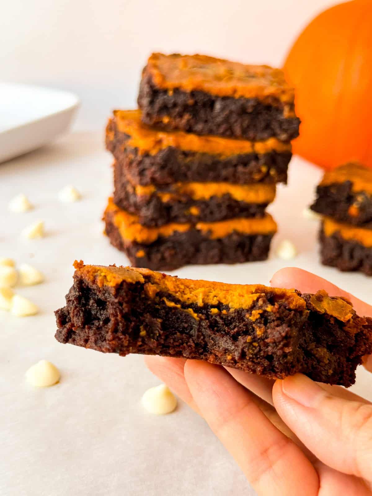 Close-up of a hand holding a slice of pumpkin cheesecake brownie showing fudgy chocolate and creamy pumpkin layers, with more brownies stacked and a pumpkin in the blurred background.