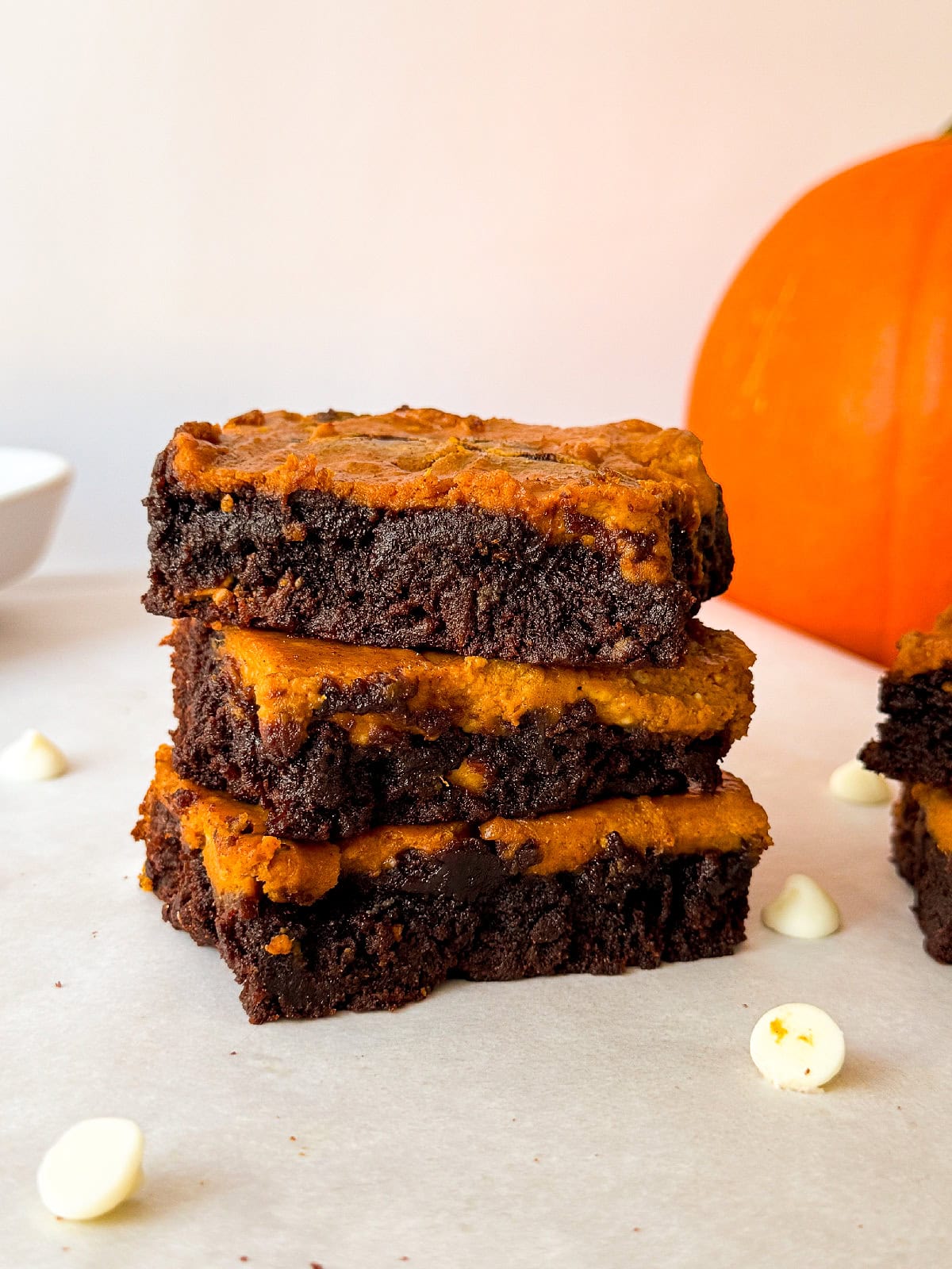 Three pumpkin cheesecake brownies stacked on parchment paper highlighting the fudgy chocolate and pumpkin layers with white chocolate chips and a pumpkin in soft focus behind.