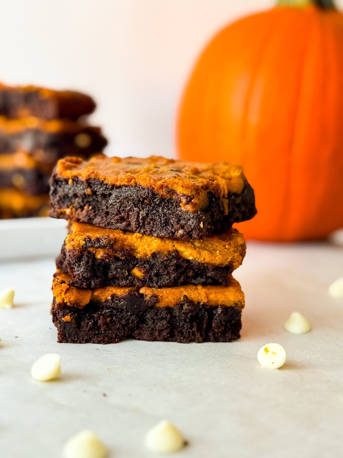 Stack of pumpkin cheesecake brownies close up showing moist chocolate brownie and bright pumpkin cheesecake layers surrounded by scattered white chocolate chips and a pumpkin in the background.