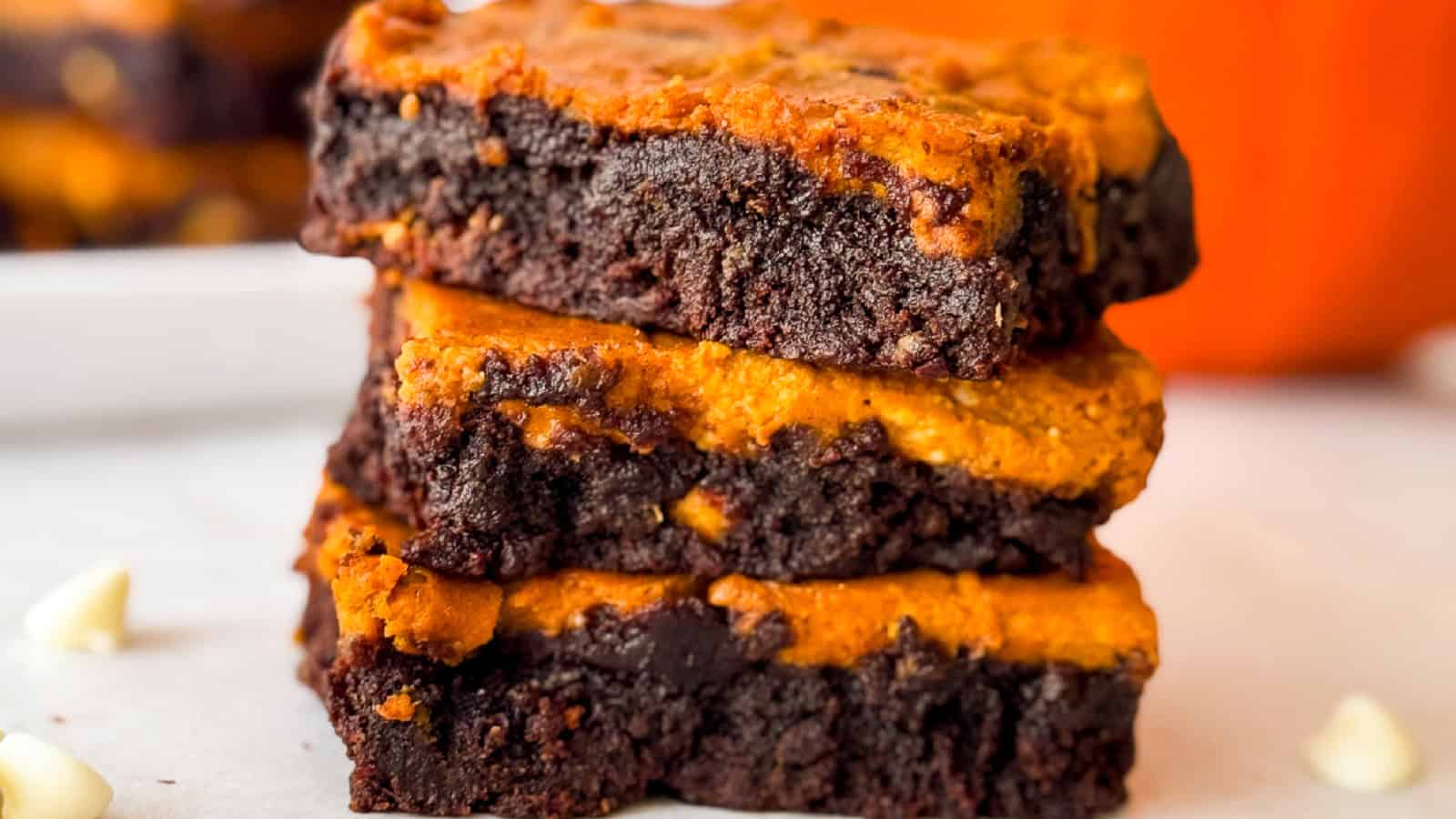 Side view of three pumpkin cheesecake brownies stacked on parchment with visible swirled chocolate and pumpkin layers, white chocolate chips, and a pumpkin in the blurred background.