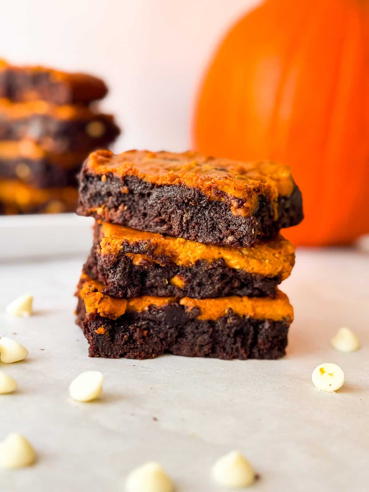 Side view of three pumpkin cheesecake brownies stacked on parchment with visible swirled chocolate and pumpkin layers, white chocolate chips, and a pumpkin in the blurred background.