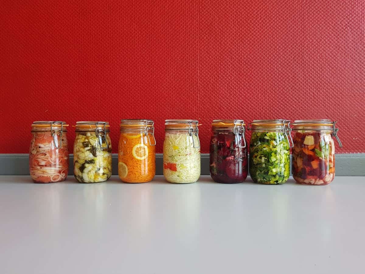 Colorful veggie pickle jars in glass containers on a white surface against a red wall.