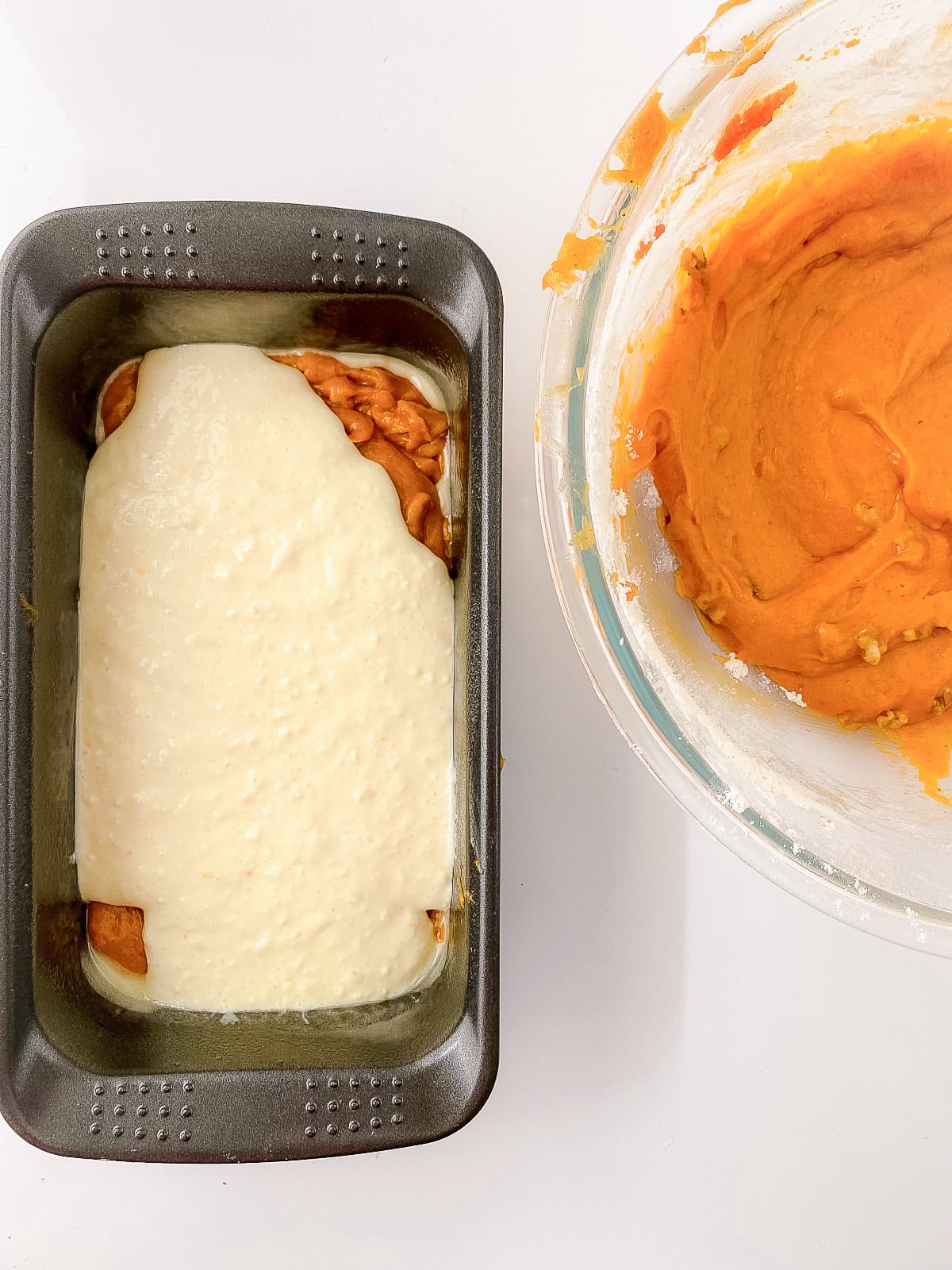 Loaf pan with first layer of pumpkin batter and cream cheese mixture poured on top, with extra pumpkin batter in a bowl beside it.
