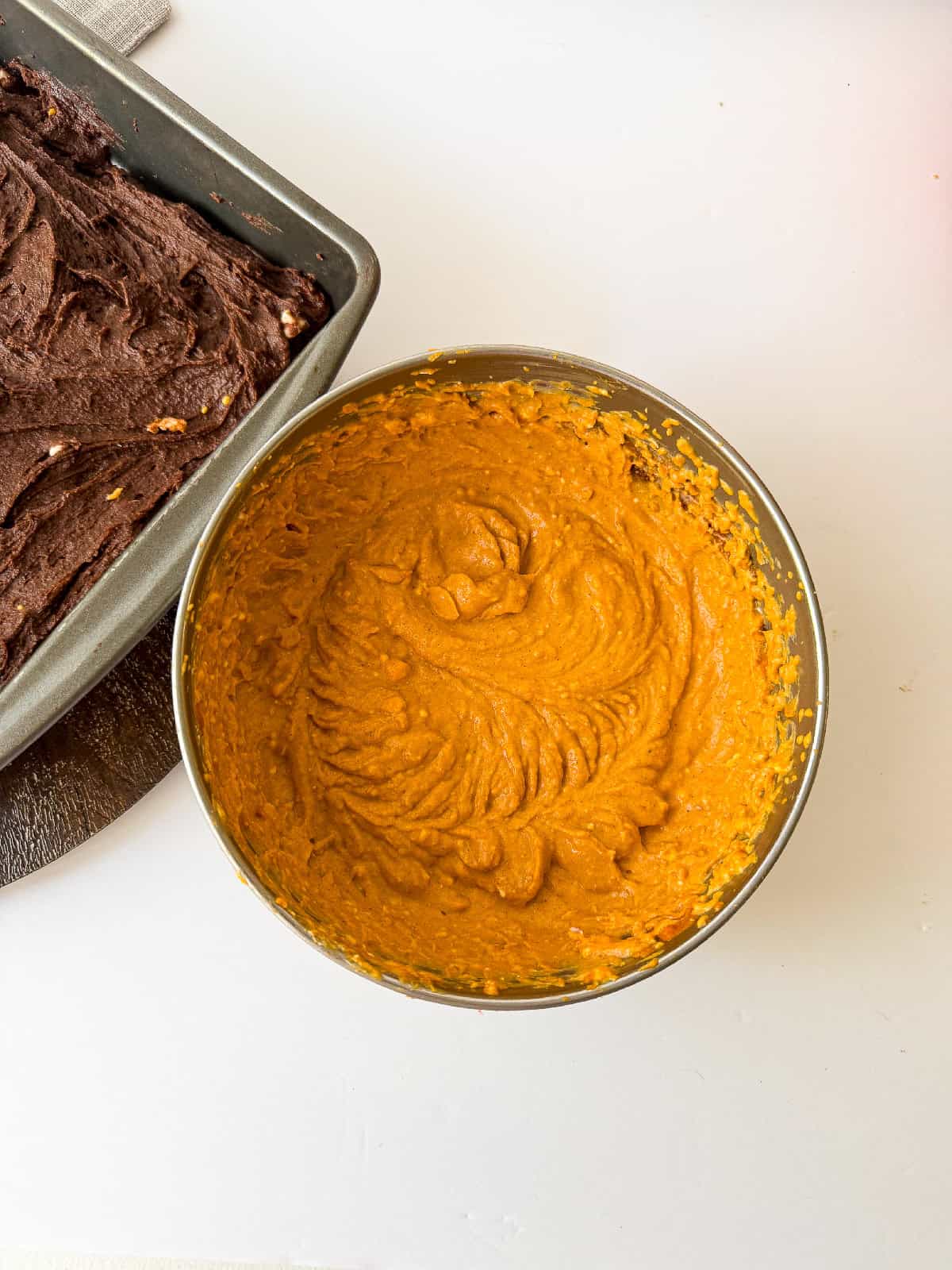 Pumpkin batter fully mixed in a bowl next to a pan of chocolate brownie batter.