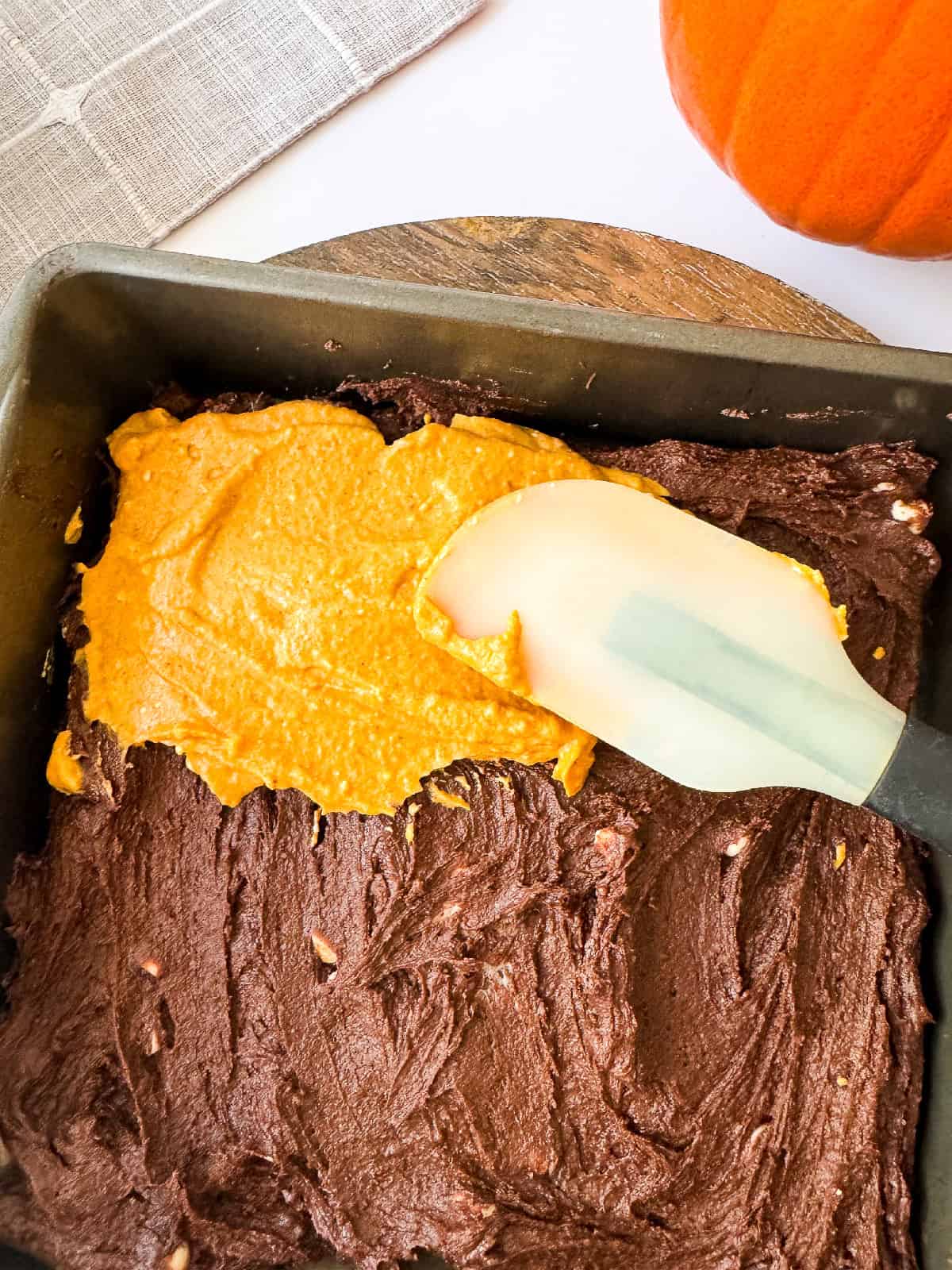 Pumpkin mixture being spread over chocolate brownie batter in a baking pan with a spatula.
