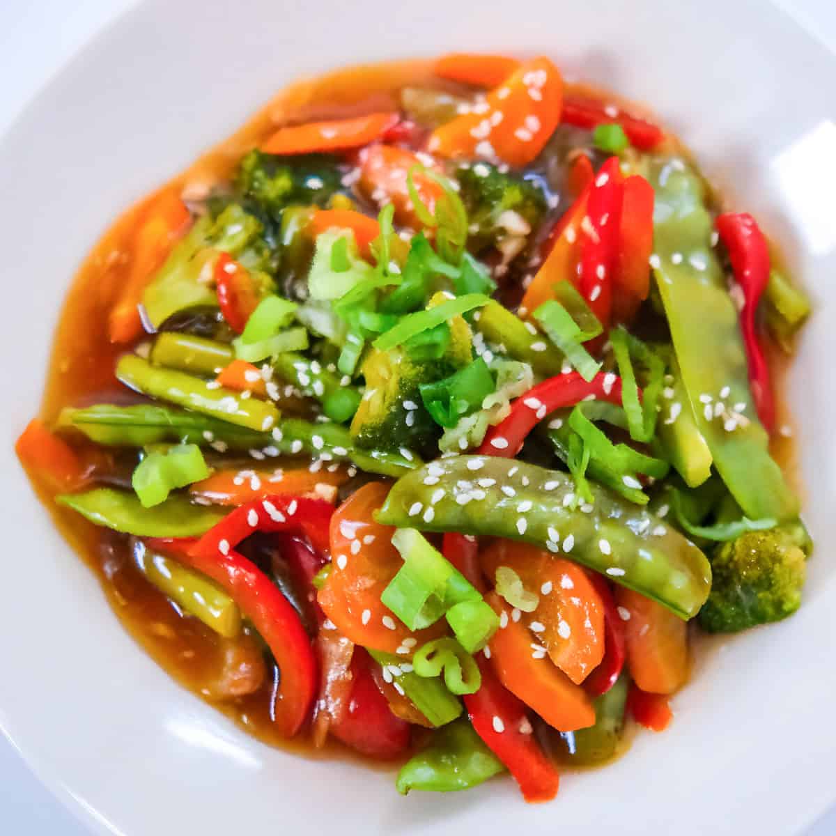 Colorful veggie teriyaki stir fry served in a white bowl with broccoli, snap peas, carrots, red bell peppers..etc and sesame seeds in a glossy sauce.