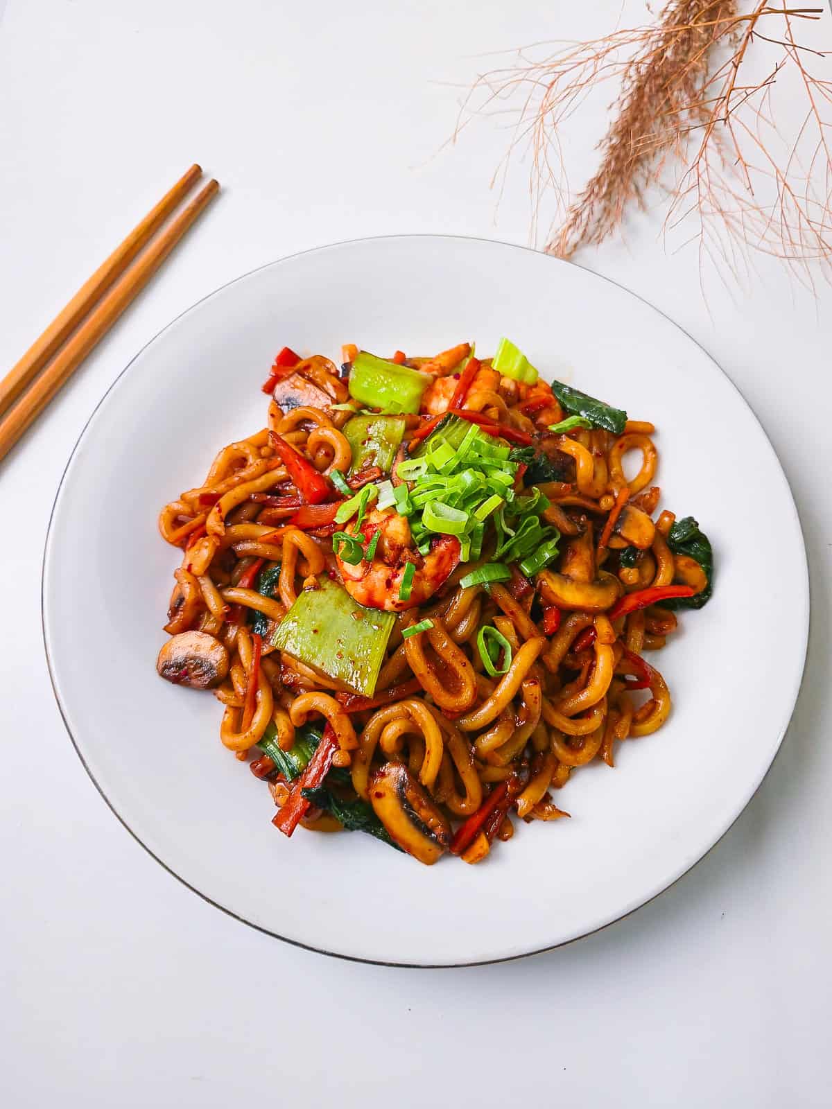 Bright overhead photo of thick udon noodles stir-fried with shrimp, bok choy, mushrooms, and colorful vegetables topped with fresh green onions.