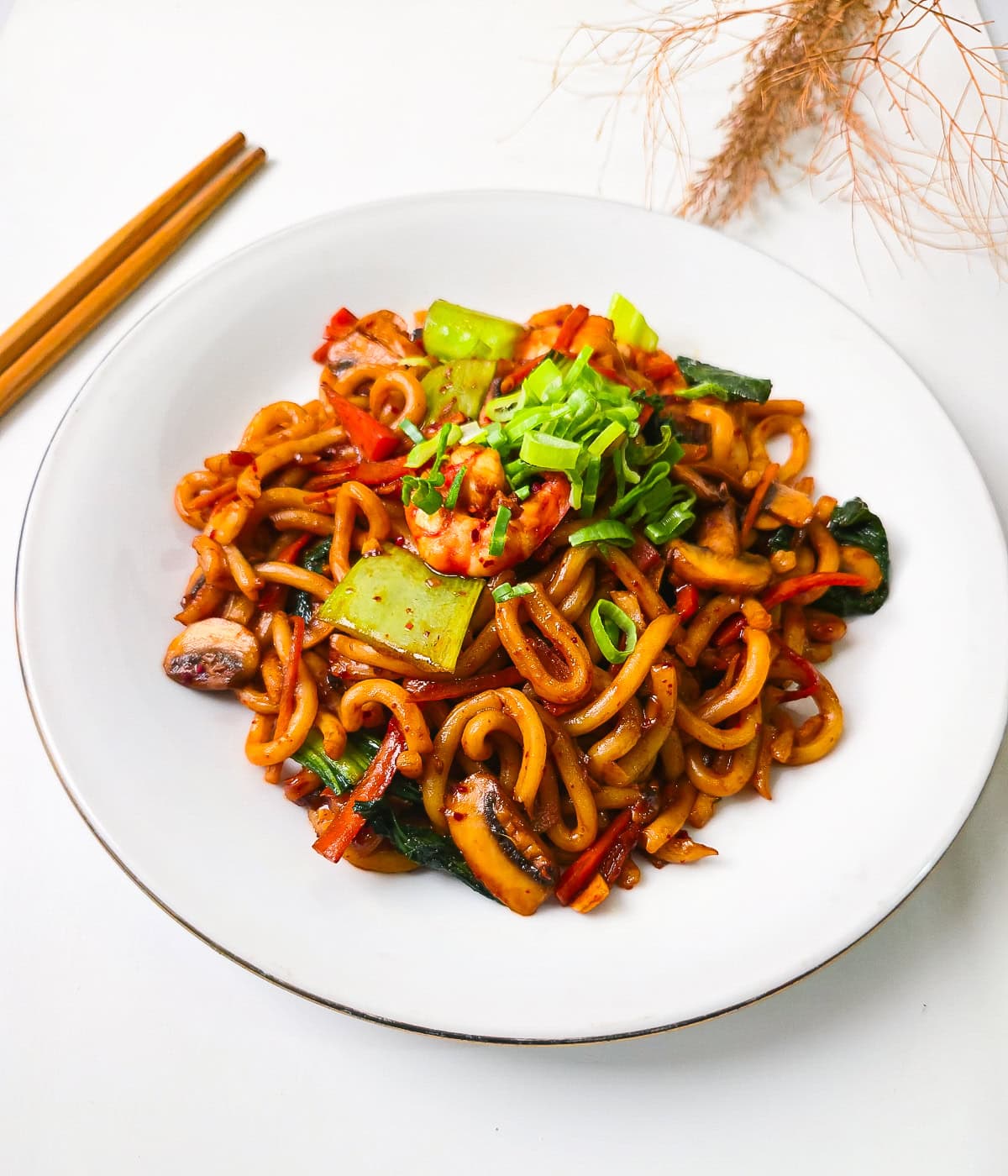 Flat lay of shrimp yaki udon noodles with vegetables and green onions on a white plate styled with dried grass and chopsticks.