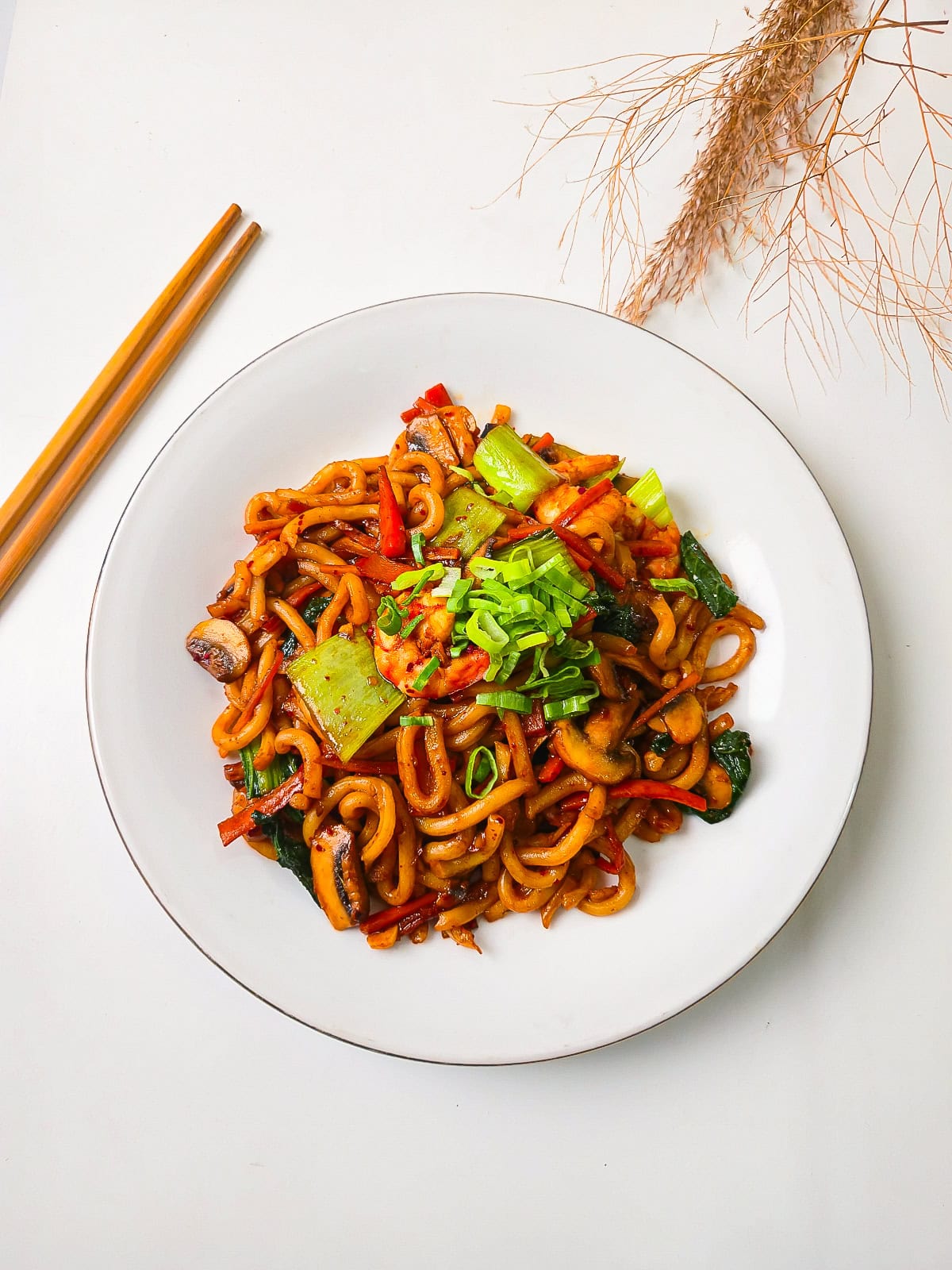 Bright overhead photo of thick udon noodles stir-fried with shrimp, bok choy, mushrooms, and colorful vegetables topped with green onions.