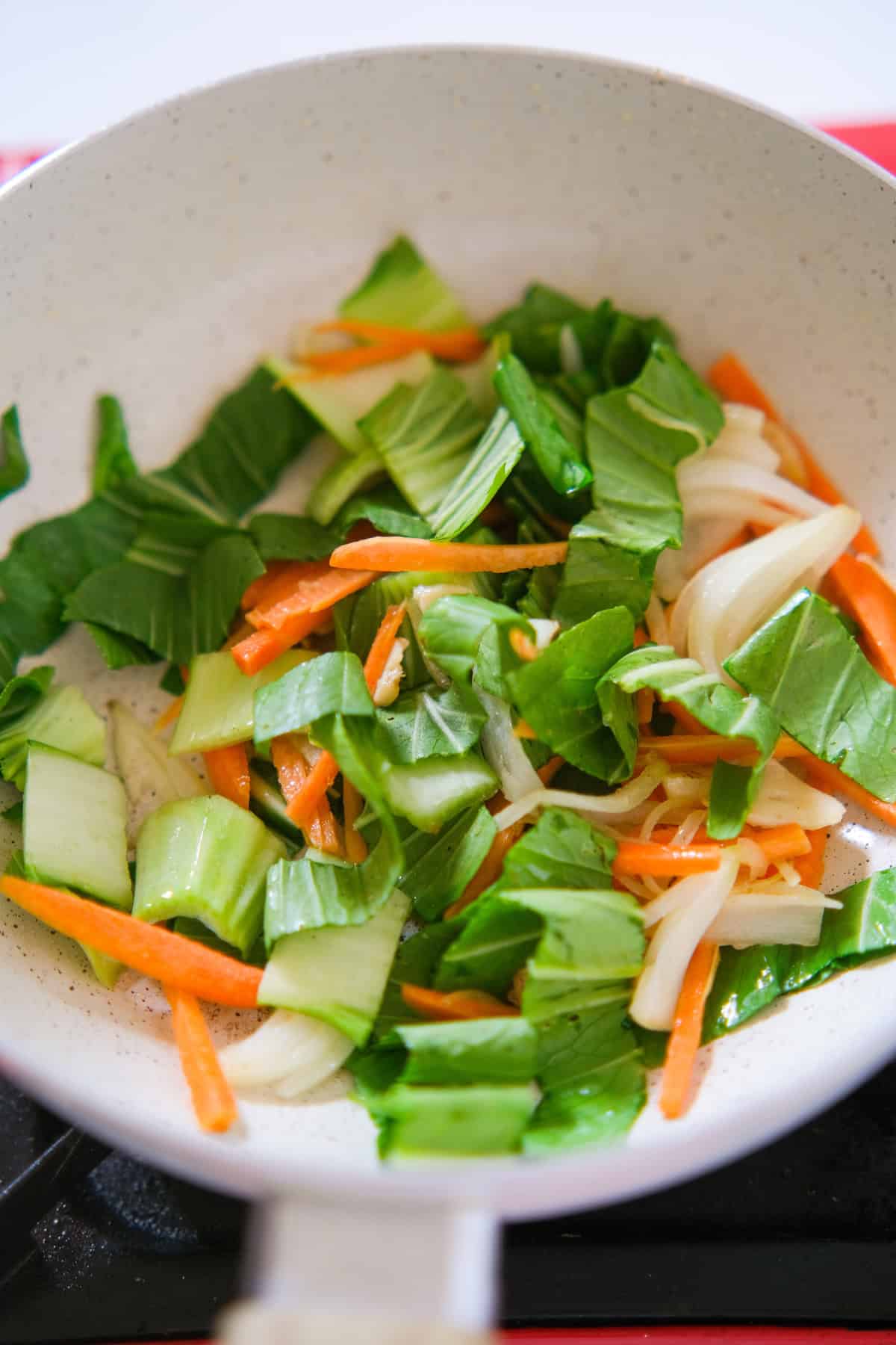 Chopped bok choy, carrots, and onions cooking in a white skillet.