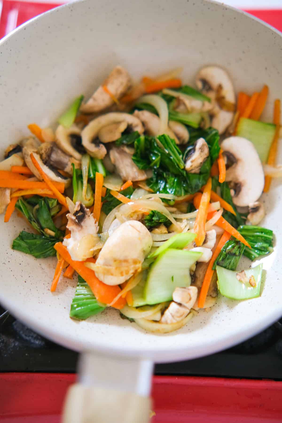 Bok choy, carrots, onions, and sliced mushrooms being stir-fried in a white skillet.