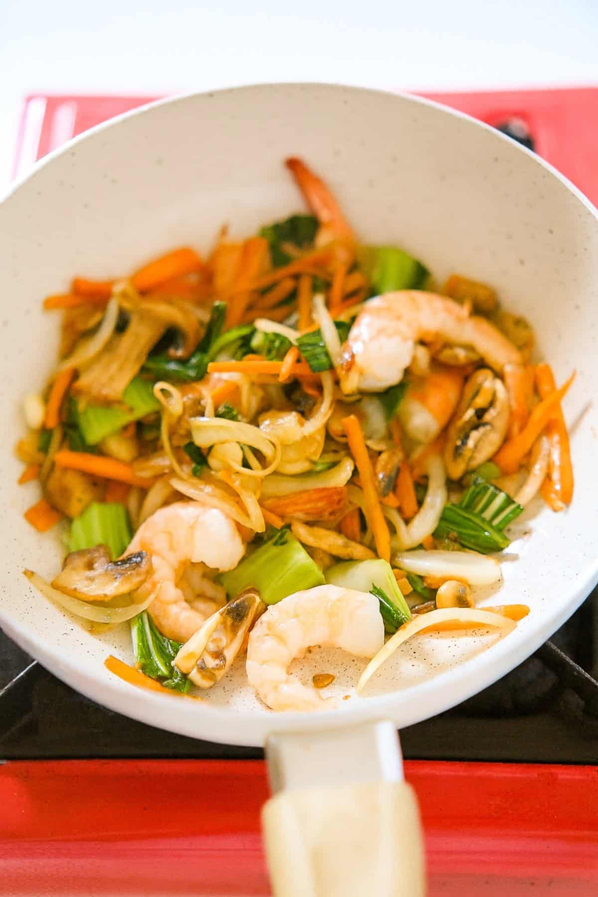 Shrimp added to the stir-fried vegetables in a white skillet on a stovetop.