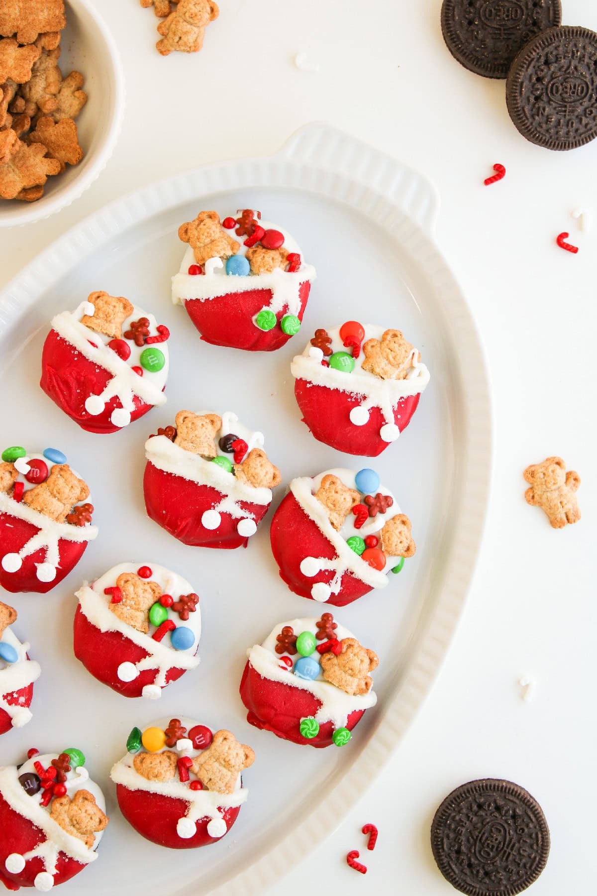 Festive Christmas Oreos arranged on a white platter with holiday sprinkles, mini candy canes, and teddy grahams.