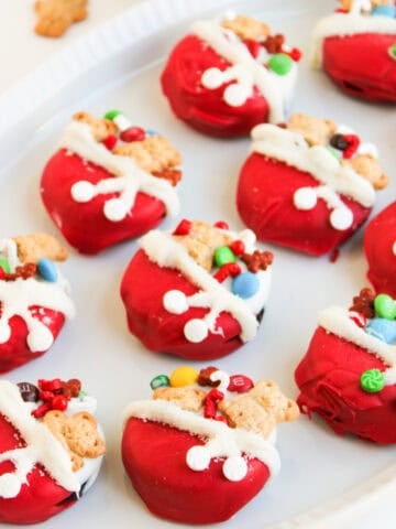 Red Santa's toy bag oreos, topped with mini teddy bear cookies, colorful candies, and white icing details, arranged neatly on a white platter.