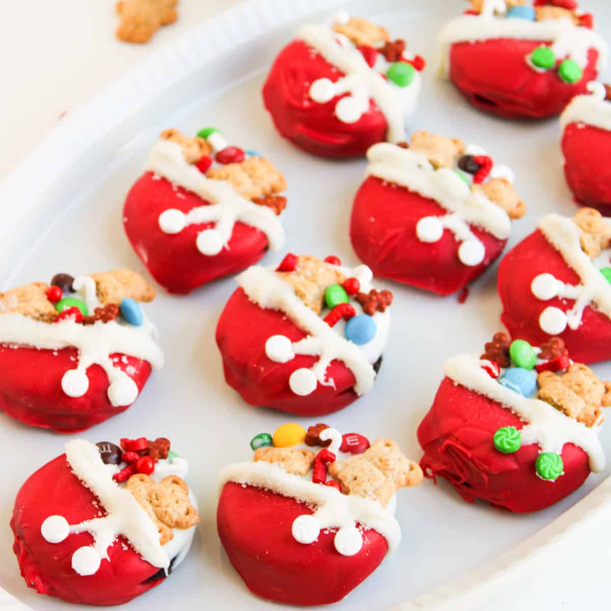 Red Santa’s toy bag oreos, topped with mini teddy bear cookies, colorful candies, and white icing details, arranged neatly on a white platter.