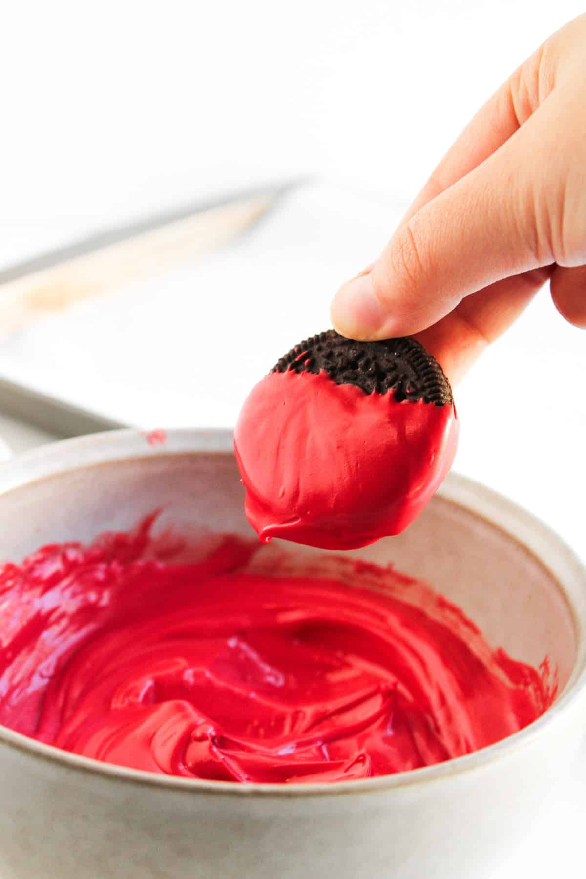 A hand dipping a chocolate sandwich cookie halfway into melted red candy coating.