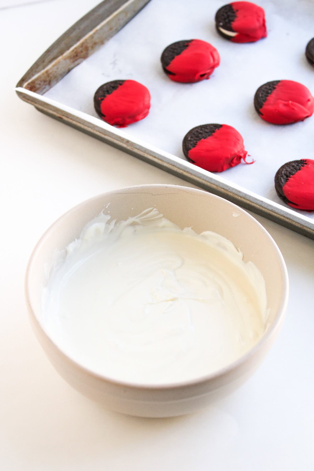 A bowl of smooth, melted white chocolate candy coating with red-dipped cookies on a tray in the background.