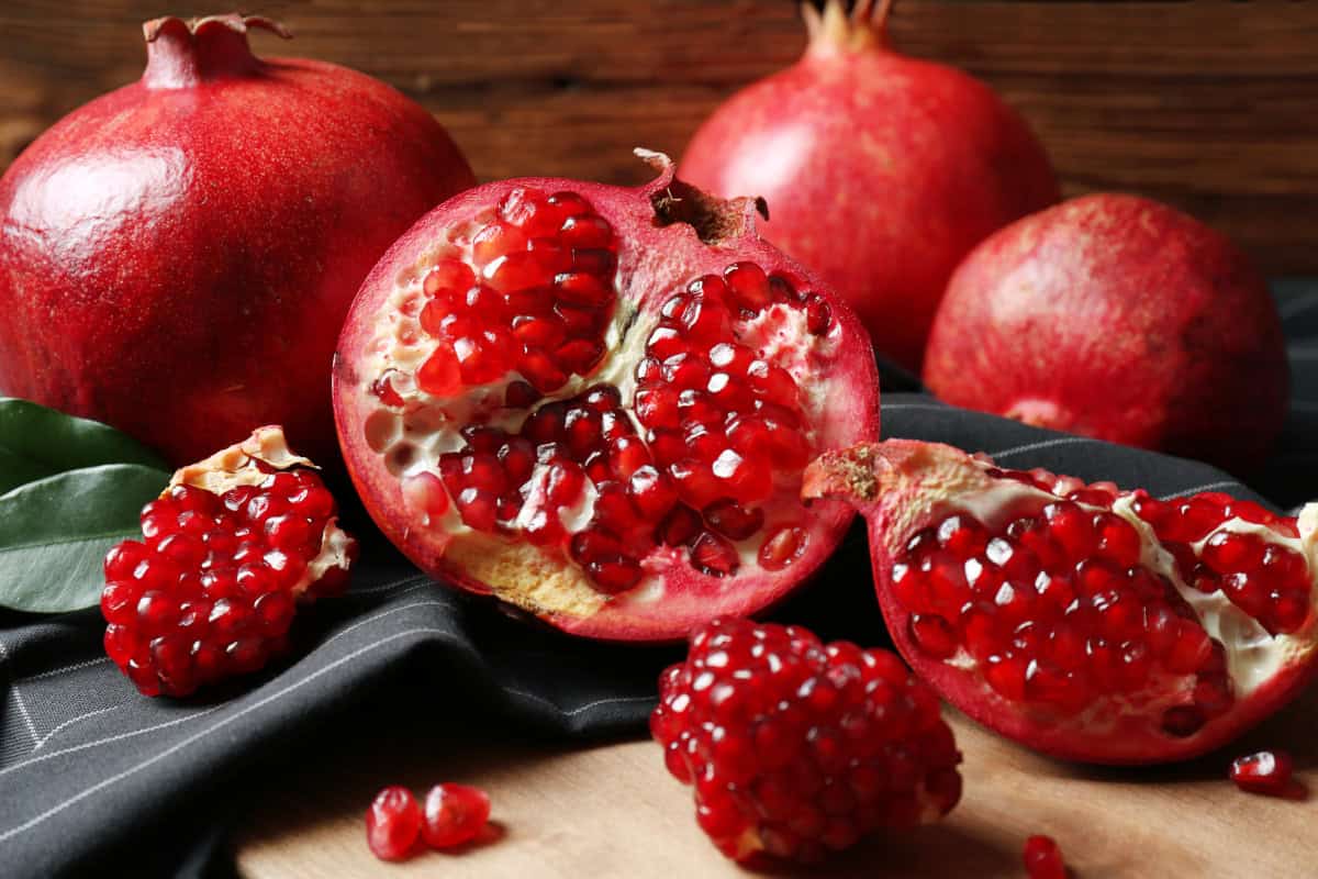 Fresh pomegranates with one cut open to reveal bright red seeds, displayed on a wooden surface with dark fabric and green leaves.