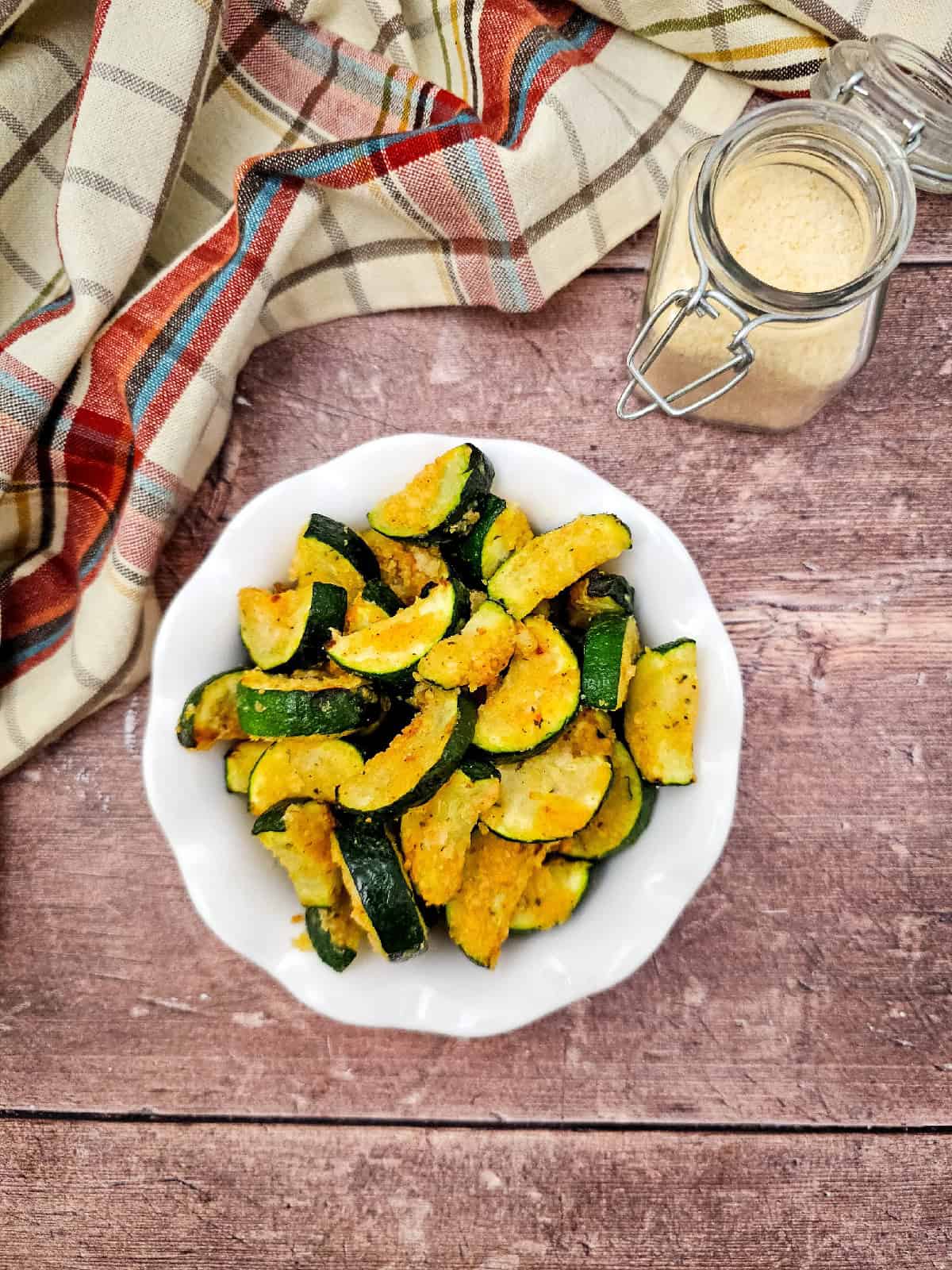 A white bowl filled with crispy air fryer zucchini parmesan wedges, set on a rustic wooden table with a jar of grated parmesan in the background and a plaid kitchen towel draped on the side.