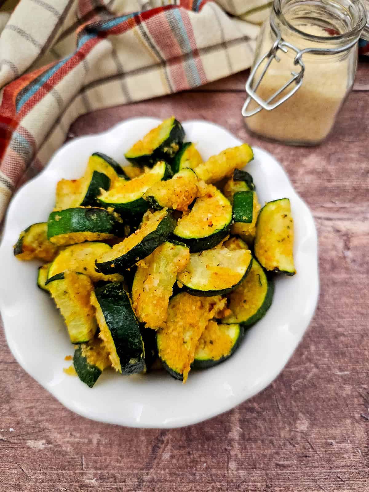Close-up view of golden, breaded air fryer zucchini parmesan pieces piled high in a white bowl, with a rustic cloth and parmesan jar in the background.