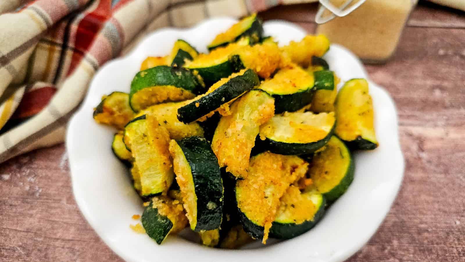 A white bowl filled with golden, breaded zucchini slices coated in vegan parmesan, set on a rustic wooden table with a plaid cloth in the background.