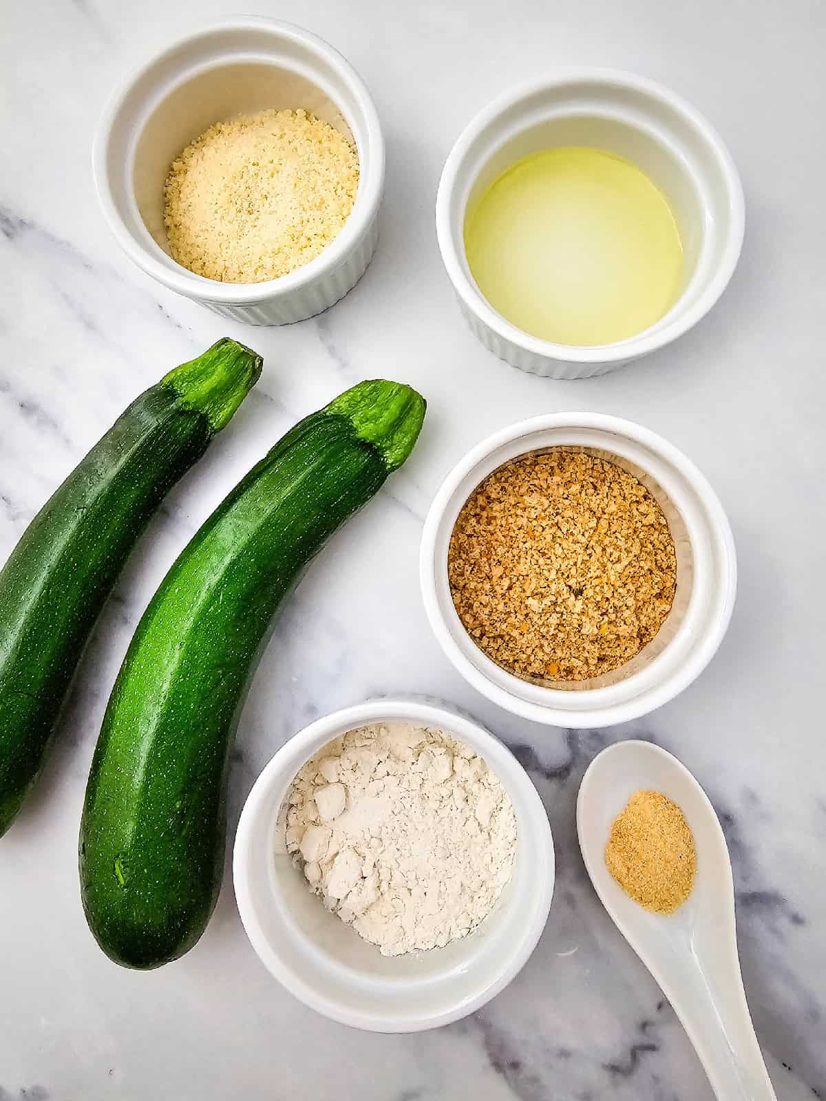 Fresh zucchini, flour, breadcrumbs, vegan grated parmesan, olive oil, and garlic powder arranged neatly in small bowls on a marble surface.