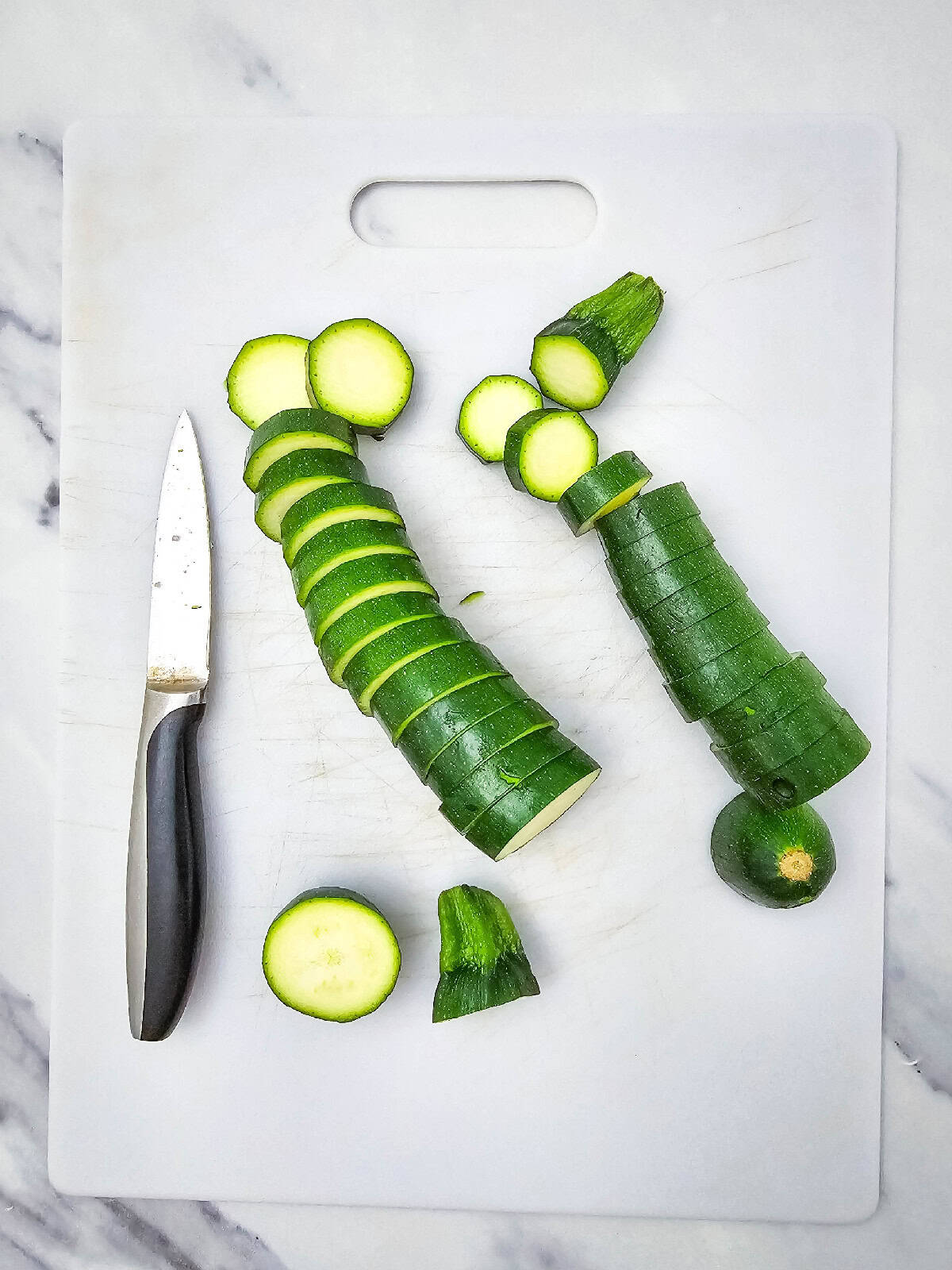 Two fresh zucchinis being sliced into even round pieces on a white cutting board with a small knife beside them.