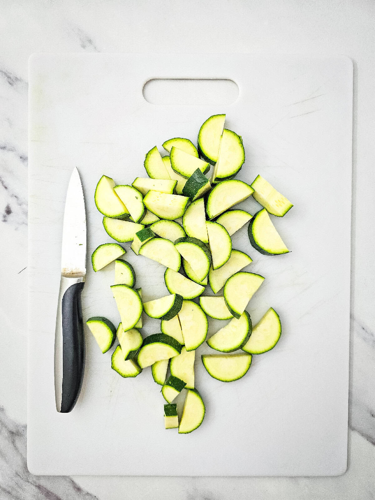 Zucchini rounds cut into halves on a white cutting board with a knife placed on the left side.