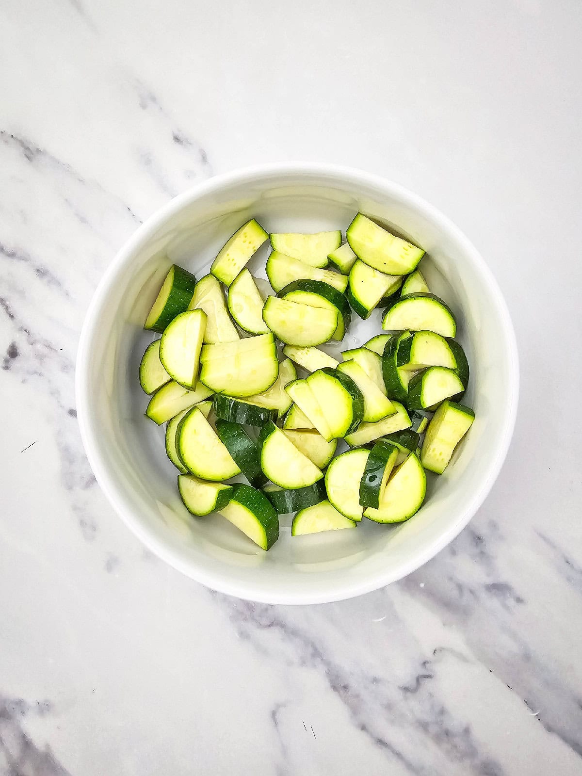 Bite-sized zucchini pieces placed in a white mixing bowl on a marble surface, ready to be seasoned.