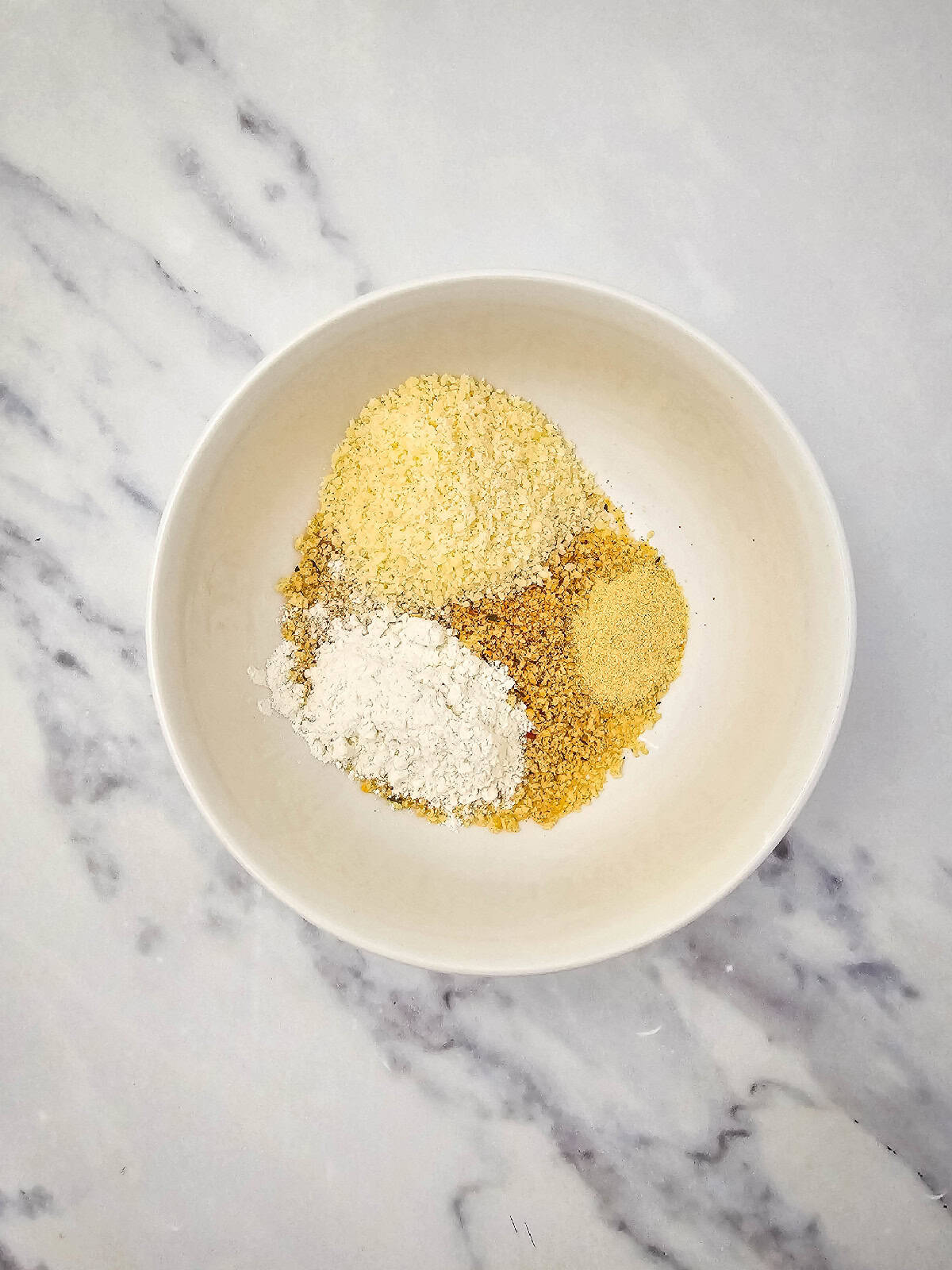 Flour, breadcrumbs, vegan grated parmesan, and garlic powder added to a white mixing bowl before mixing.