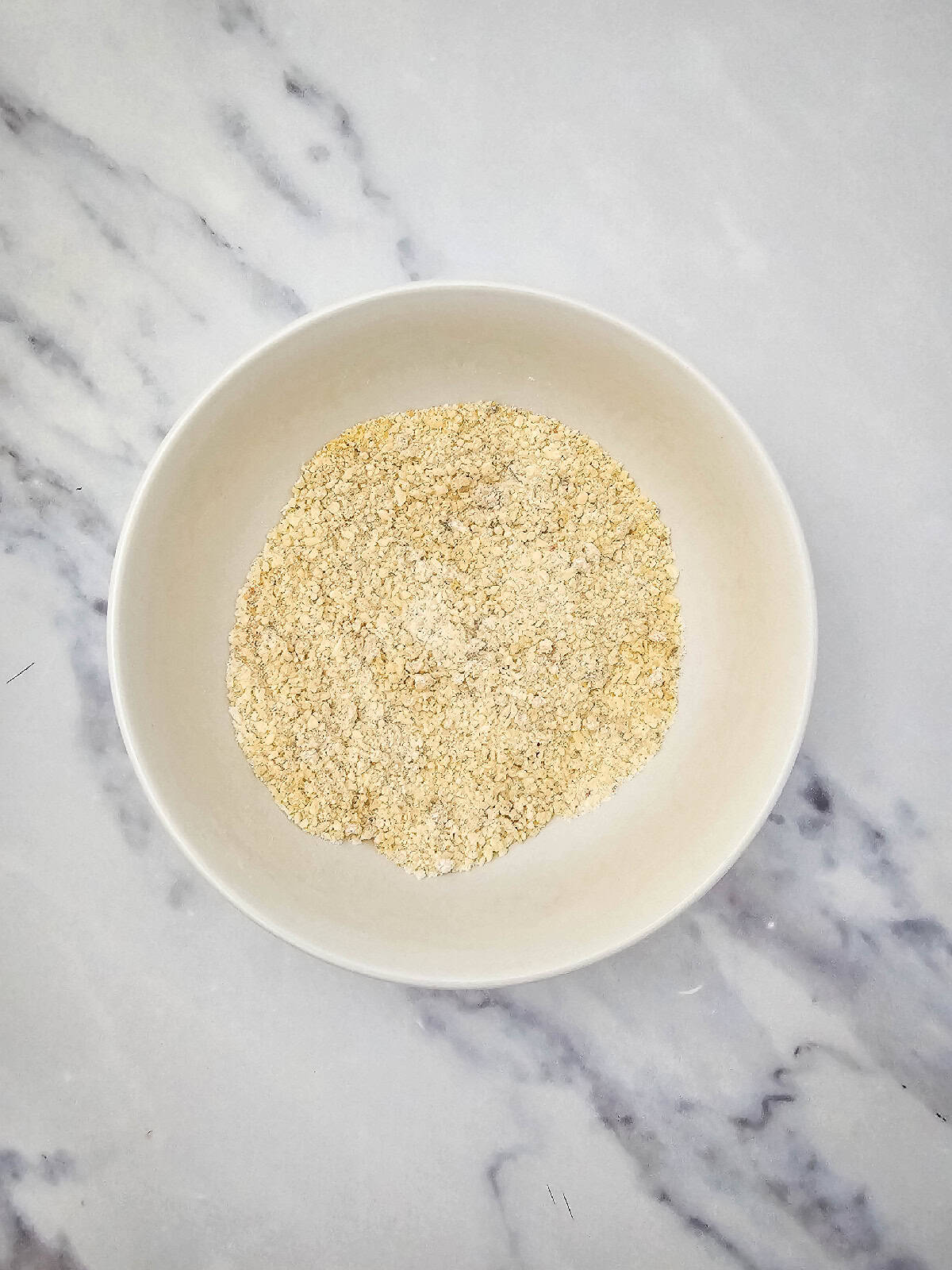 Breadcrumb and vegan parmesan mixture fully combined in a white mixing bowl on a marble countertop.