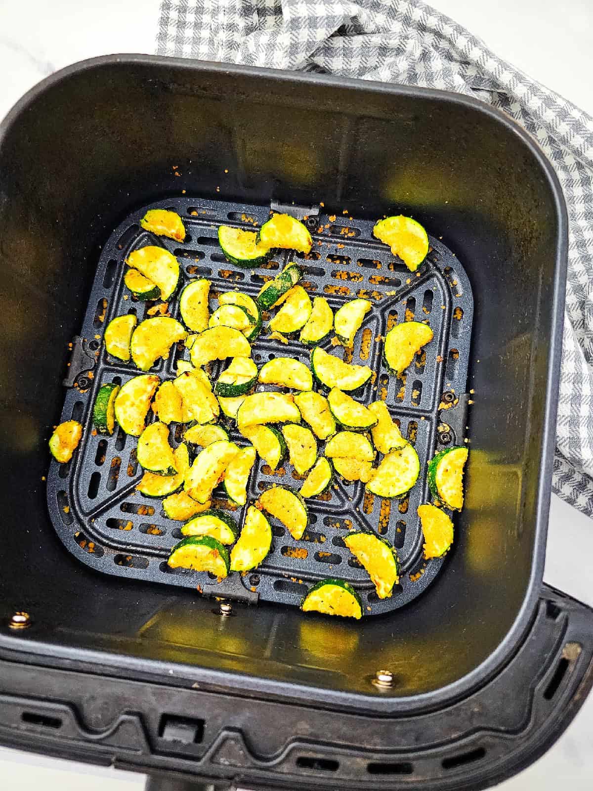 Golden zucchini slices in the air fryer basket halfway through cooking, with a checkered kitchen towel on the side.