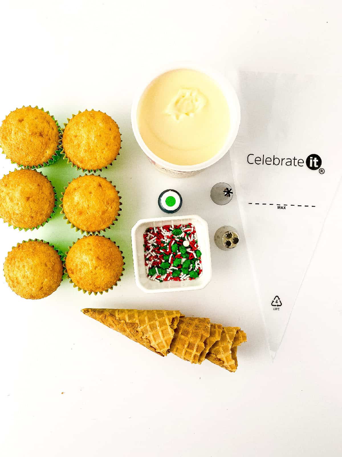 Ingredients for making Christmas tree cupcakes, including vanilla cupcakes, white frosting, holiday sprinkles, green food coloring, waffle cones, piping tips, and a piping bag arranged on a white background.