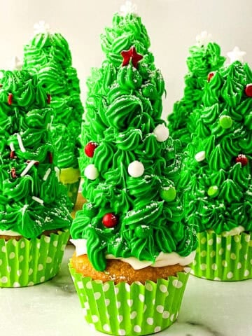 Festive Christmas tree cupcakes decorated with bright green frosting, white sprinkles, and red star toppers, arranged on a white surface.