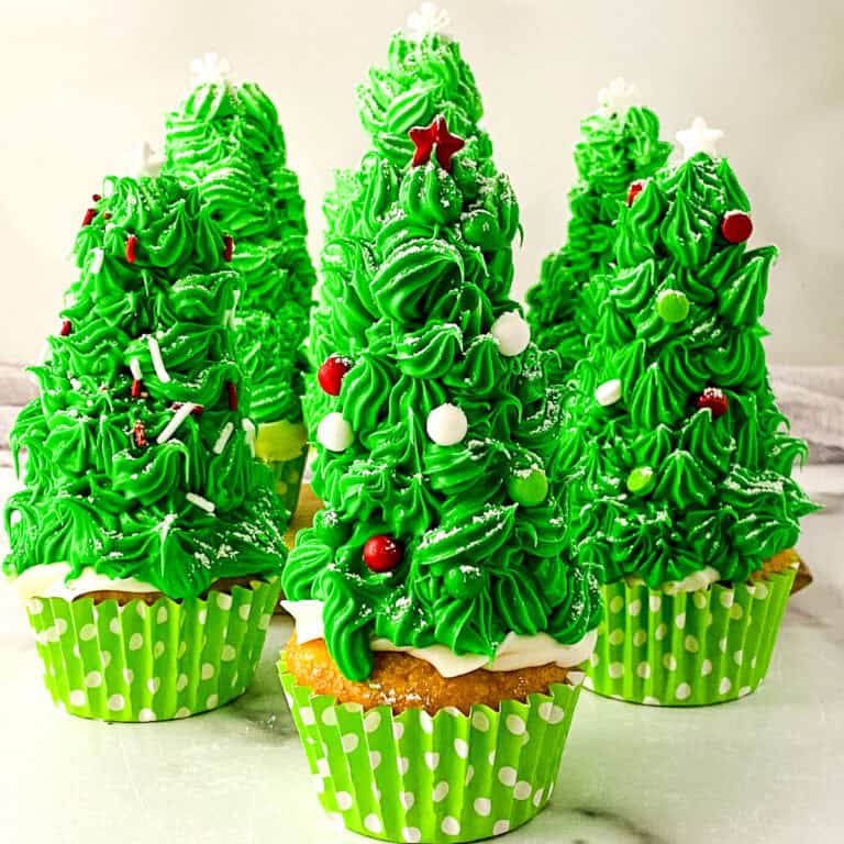 Festive Christmas tree cupcakes decorated with bright green frosting, white sprinkles, and red star toppers, arranged on a white surface.
