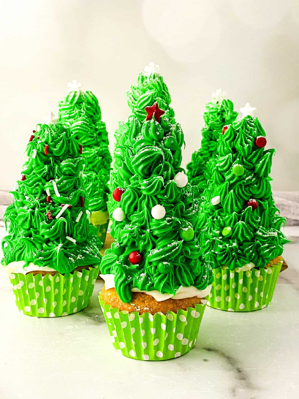 Christmas tree cupcakes decorated with bright green frosting, white snowflake toppers, red and white sprinkles, and sugar dusting, arranged in green polka dot liners.