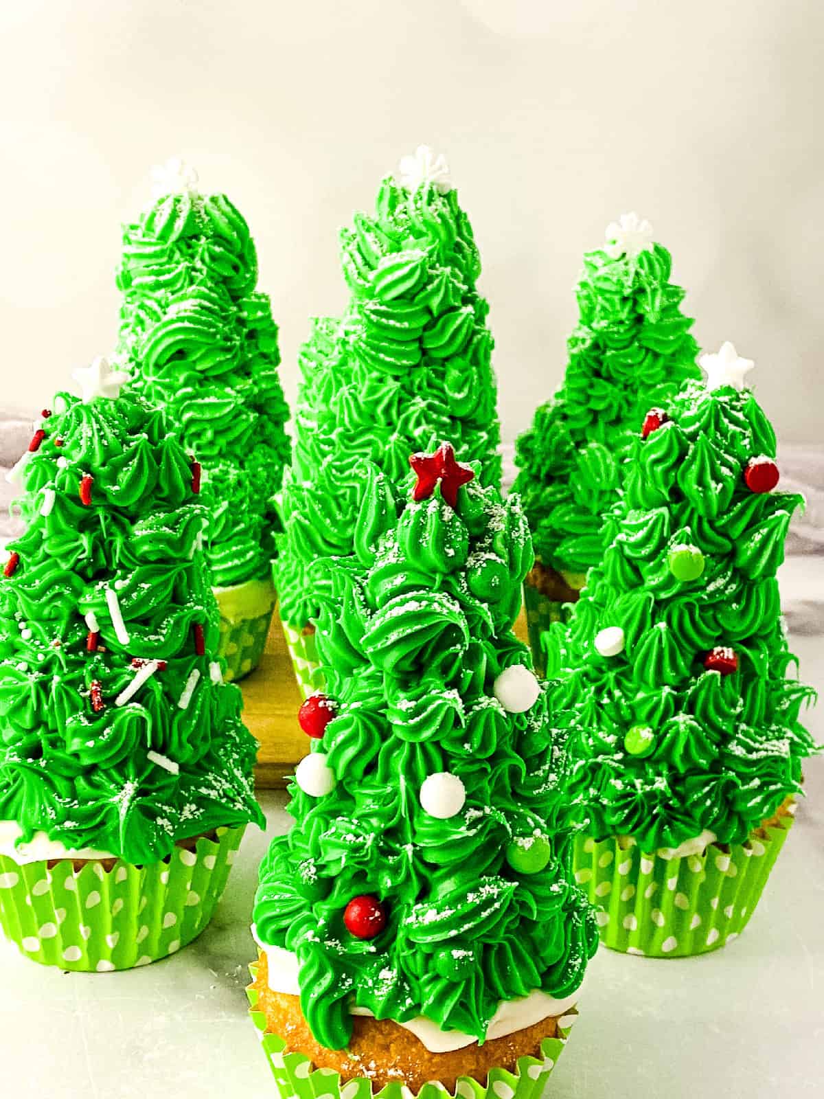Festive Christmas tree cupcakes with green buttercream frosting, red star toppers, and white sprinkles arranged in a group on a light surface.