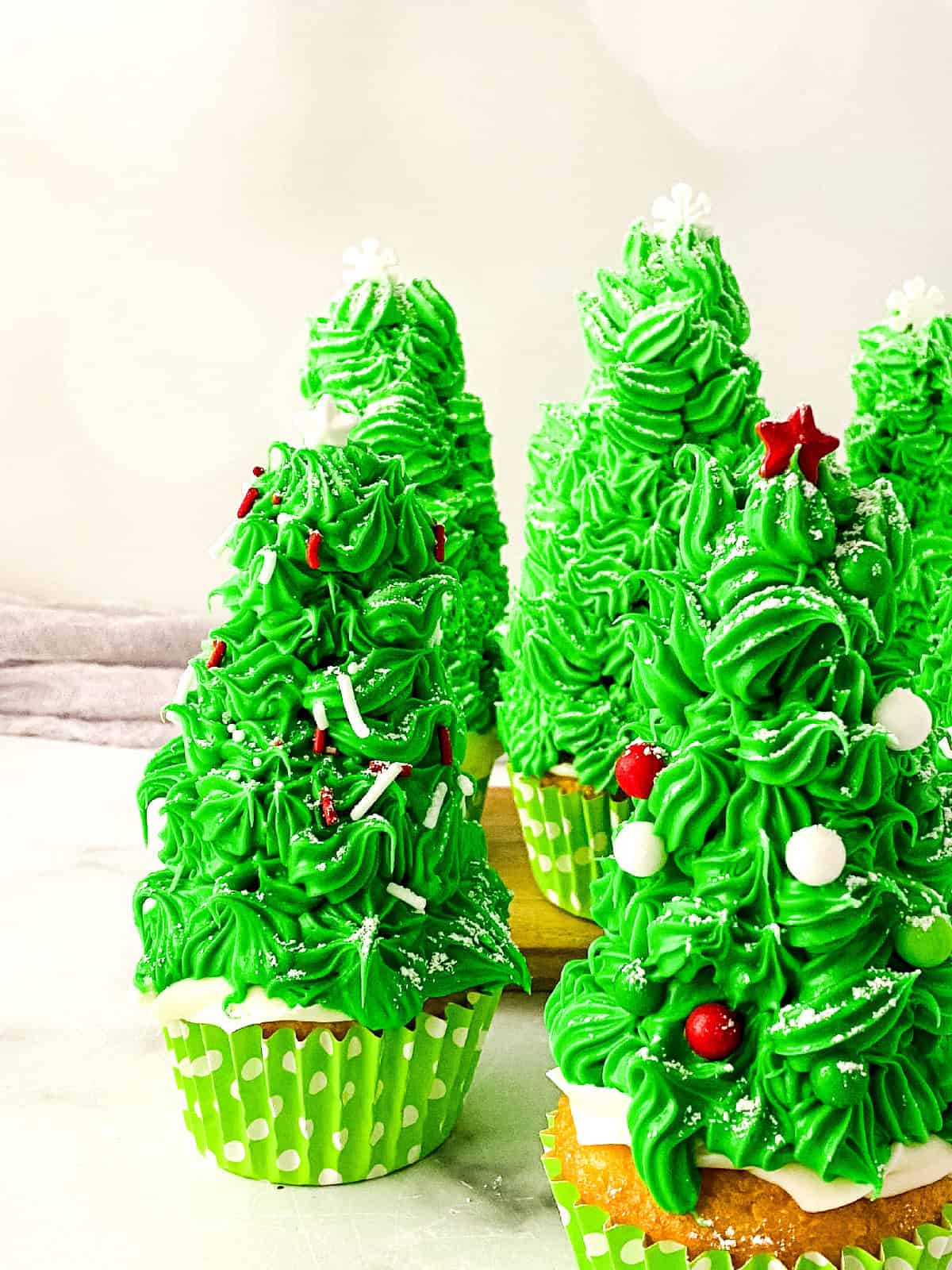 Close-up of Christmas tree cupcakes decorated with piped green frosting, red and white ornaments, and snowflake sprinkles.