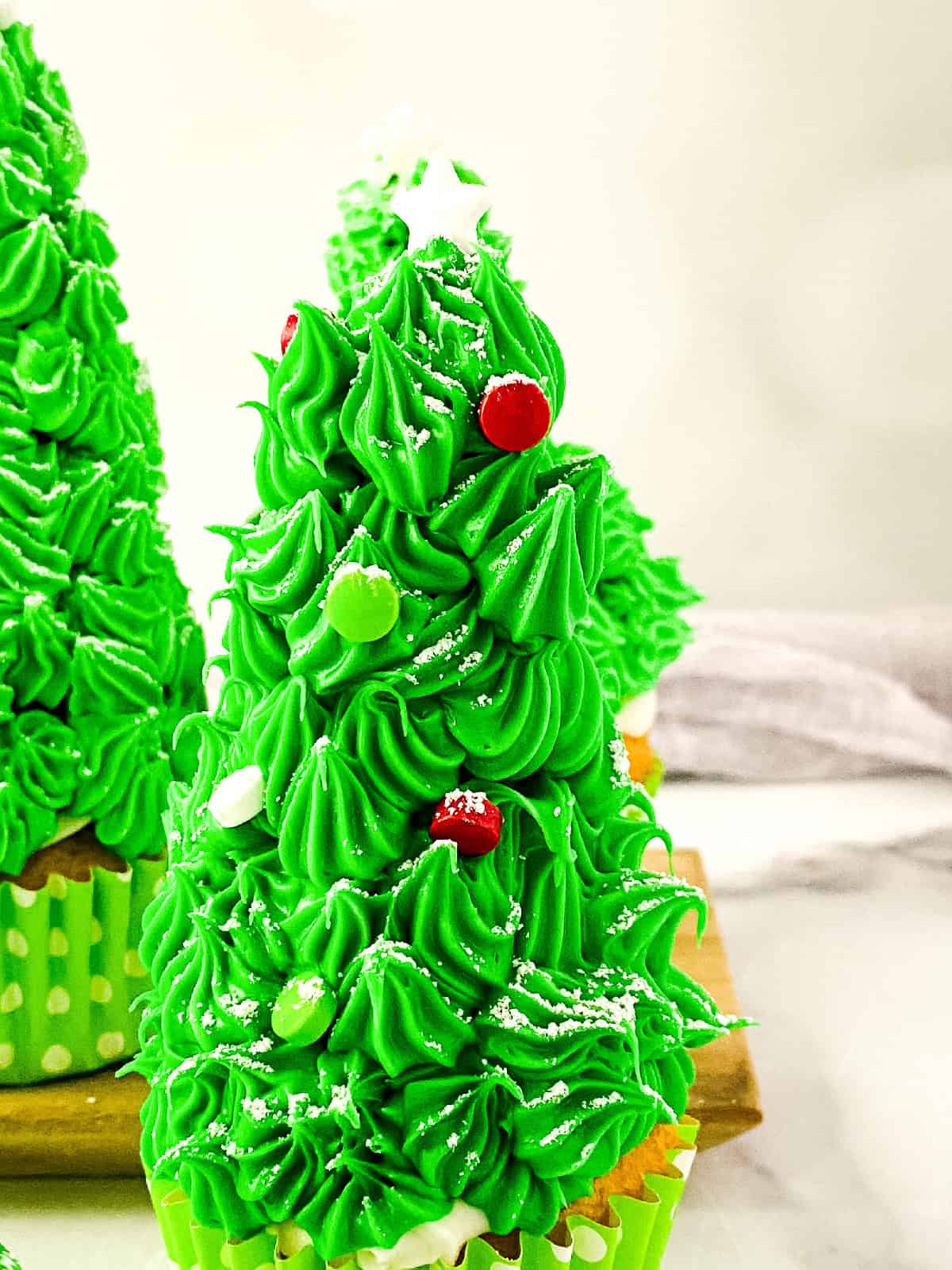 Single Christmas tree cupcake decorated with piped green frosting, red and green candies, and a white star topper, with more cupcakes in the background.
