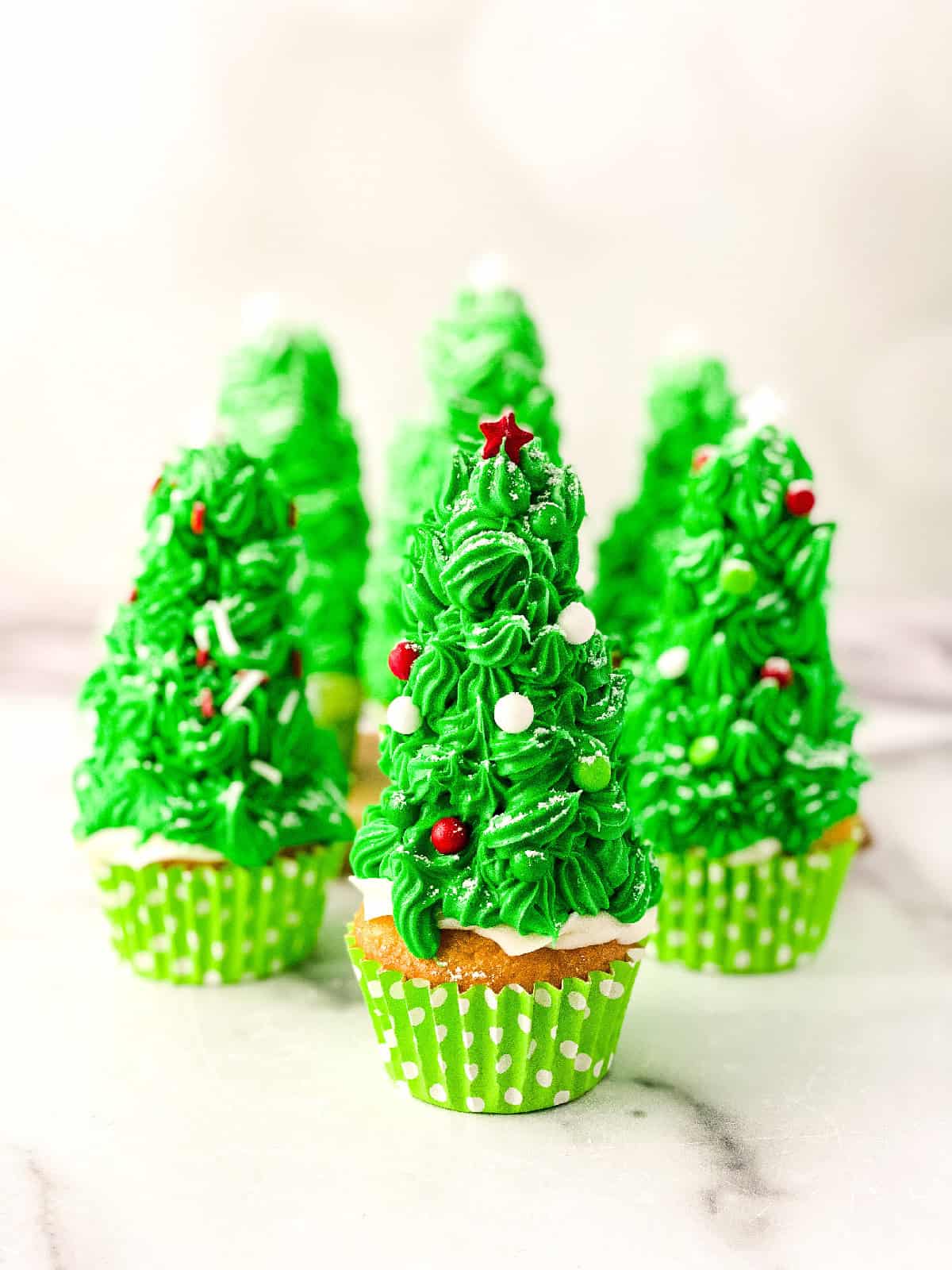 Front view of multiple Christmas tree cupcakes with green frosting, candy ornaments, and powdered sugar snow, arranged on a marble surface.
