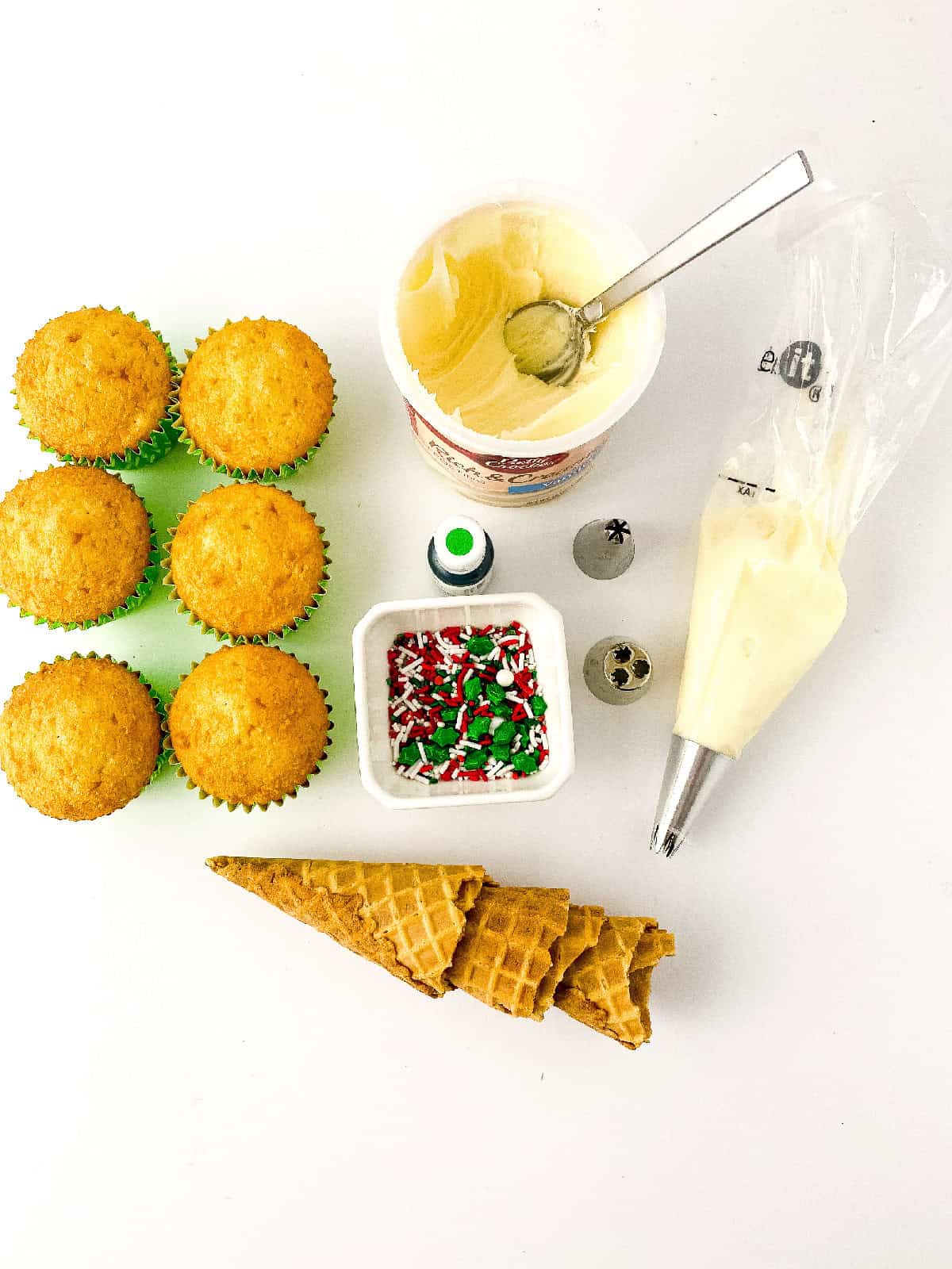 Ingredients for making Christmas tree cupcakes, including vanilla cupcakes, white frosting, piping tips, green food coloring, waffle cones, and holiday sprinkles arranged on a white background.