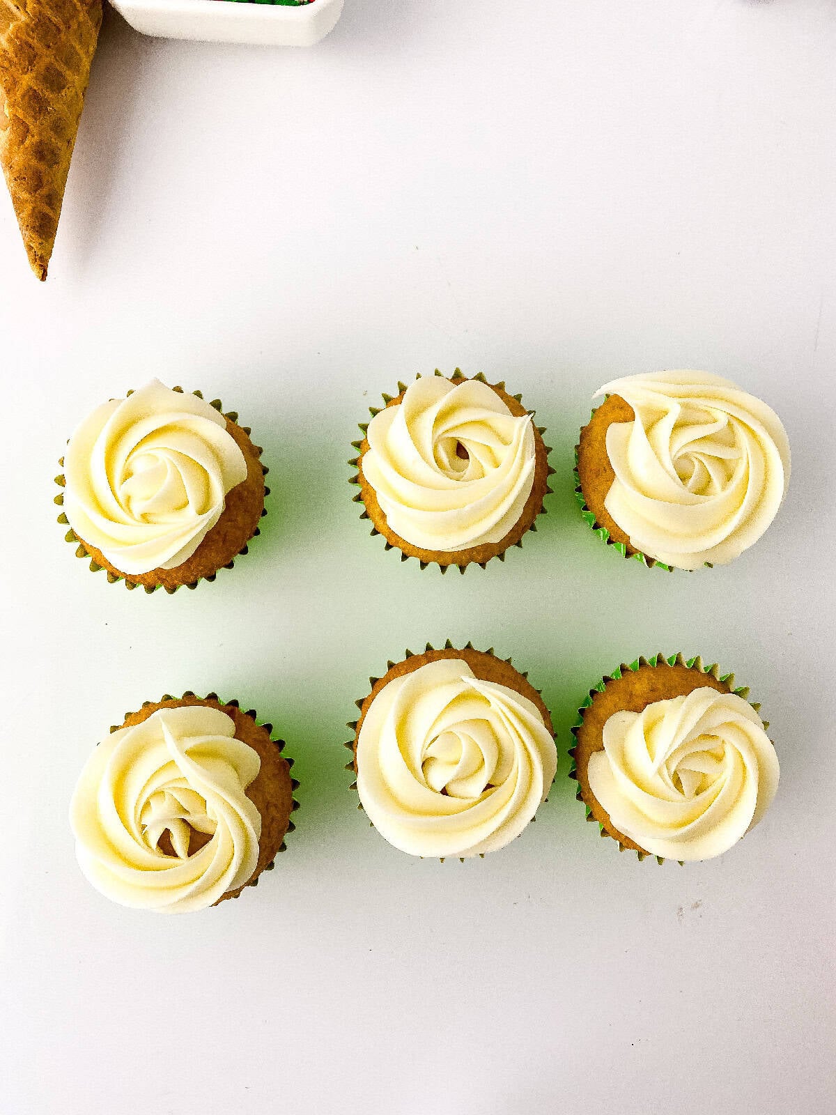 Six cupcakes topped with white buttercream rosettes, arranged neatly on a white surface with decorating tools nearby.