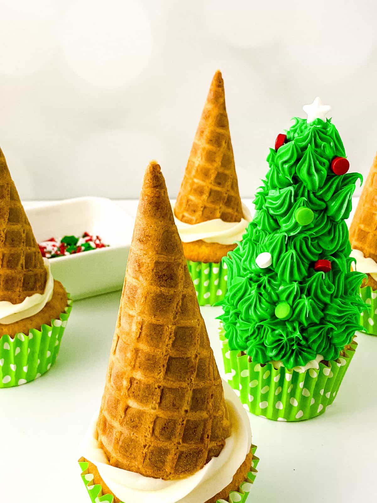 One cupcake decorated as a Christmas tree with green frosting and red and white sprinkles, surrounded by undecorated cone-topped cupcakes.