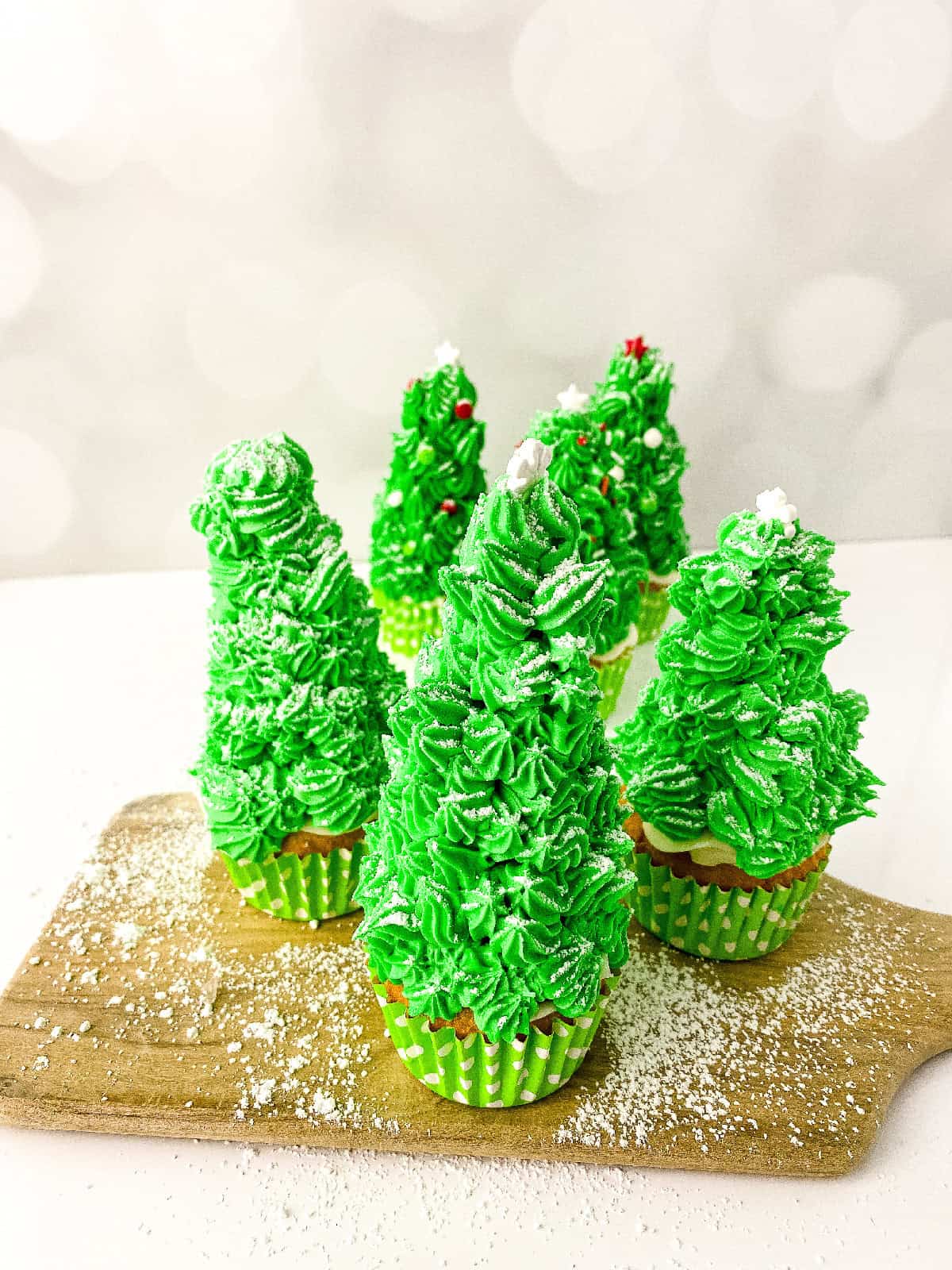 Finished Christmas tree cupcakes with green frosting, white star toppers, and powdered sugar snow, arranged on a wooden board.