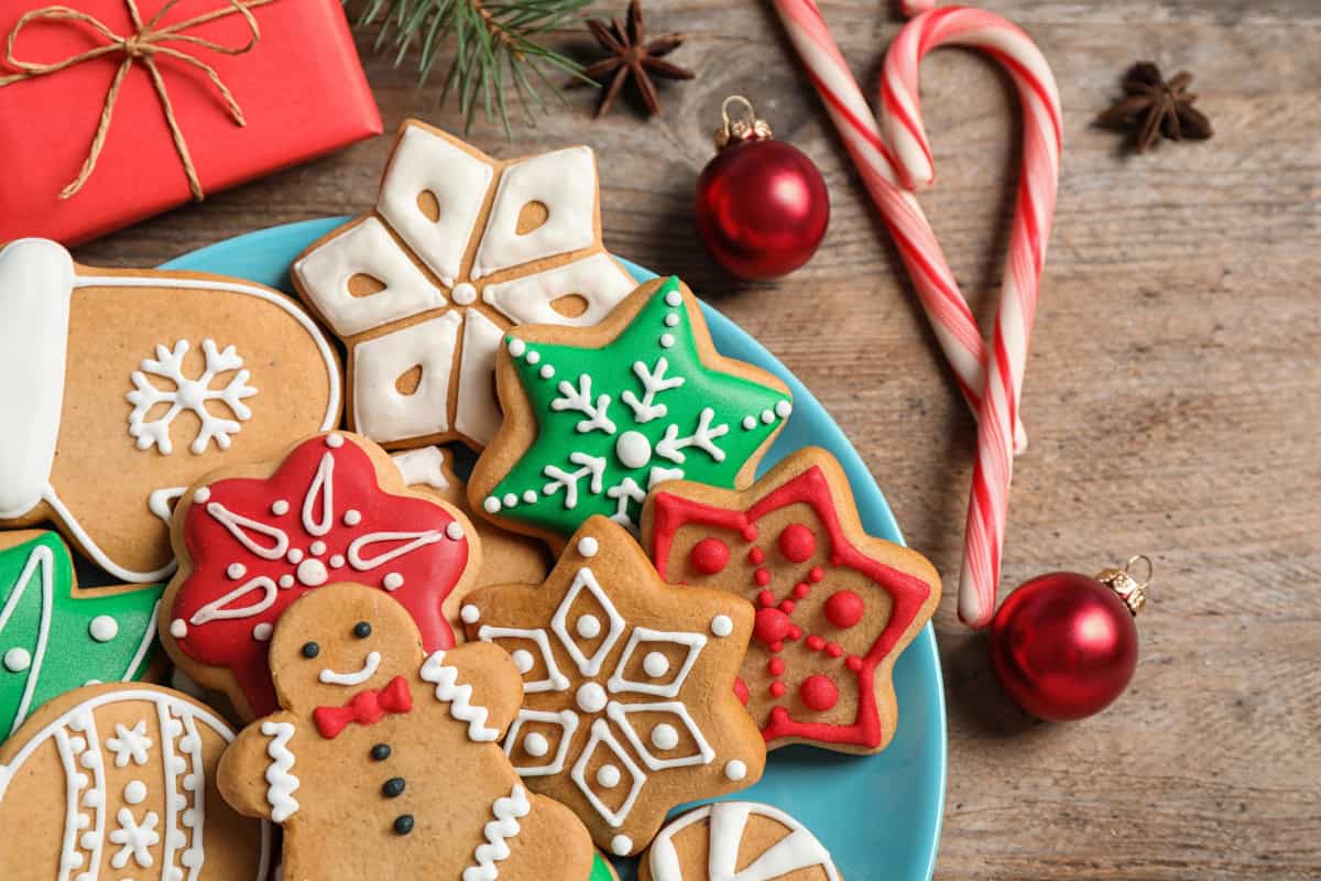 Festive holiday sugar cookies decorated with colorful royal icing, surrounded by candy canes, red ornaments, and wrapped gifts on a wooden table.