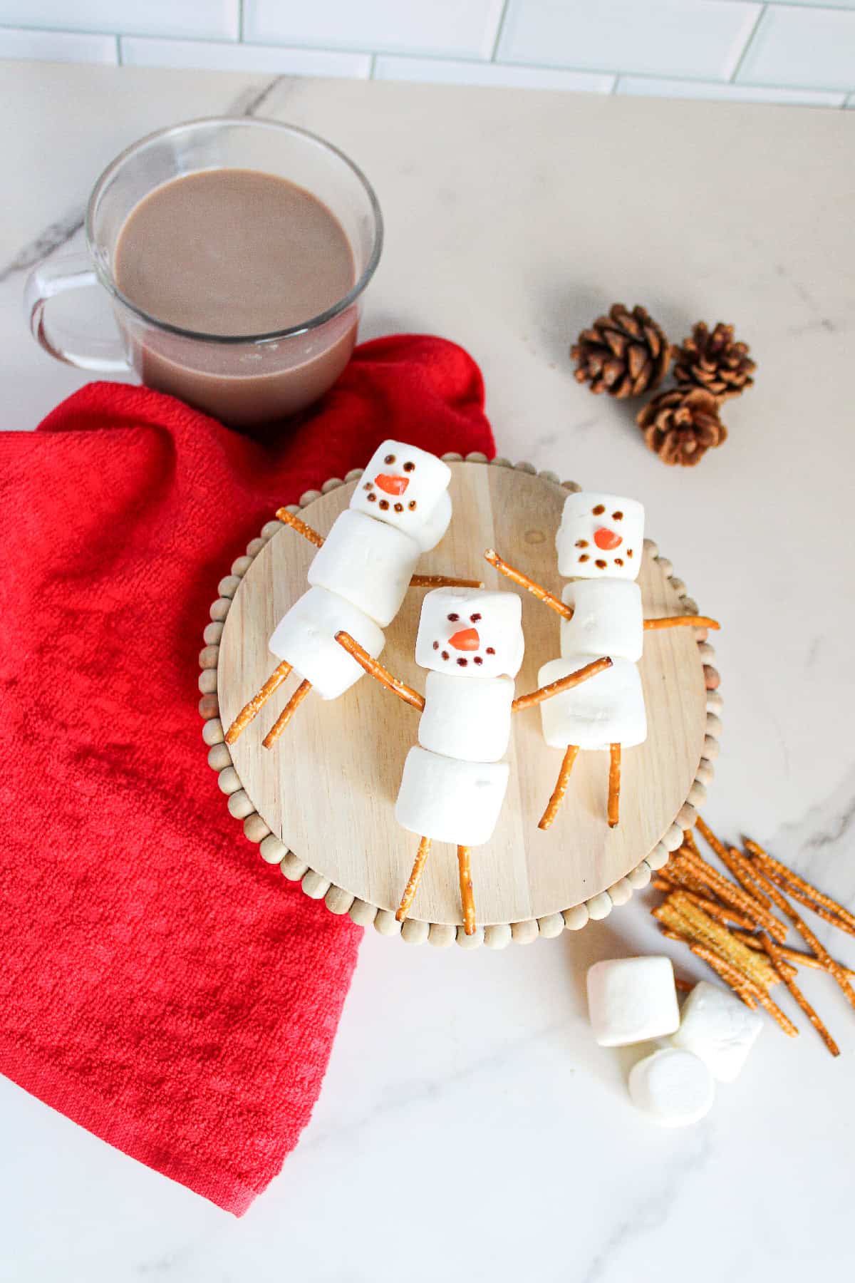 Three marshmallow snowmen with pretzel arms and legs arranged on a round wooden tray with a red cloth, hot chocolate, and pinecones.